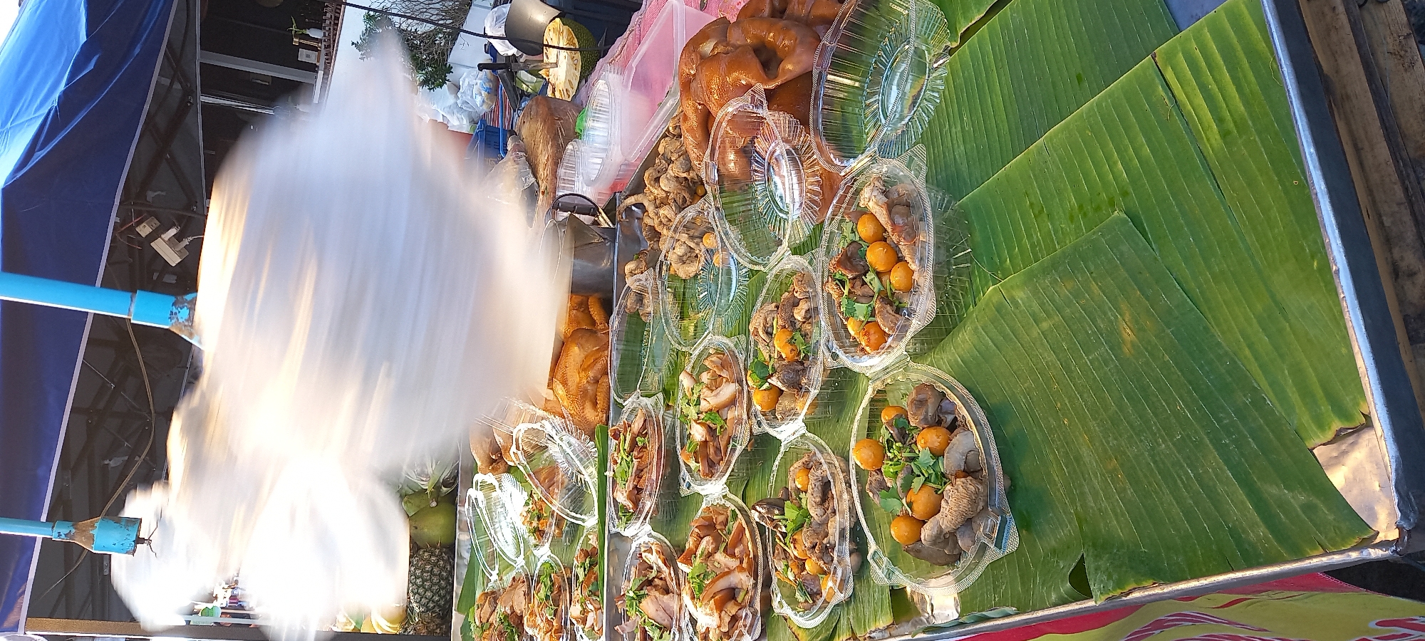 This image depicts a street food vendor in Thong Sala, Thailand. The vendor's stall features a variety of prepared foods in clear plastic containers, arranged on a surface lined with green banana leaves. The food items appear to include braised pork, chicken, and eggs. In the background are other items of street food, possibly jackfruit. Also, the shot includes a white blur in the top-left corner of the image, possibly a reflection or lighting effect. The overall scene suggests a bustling street food environment in a Southeast Asian location.