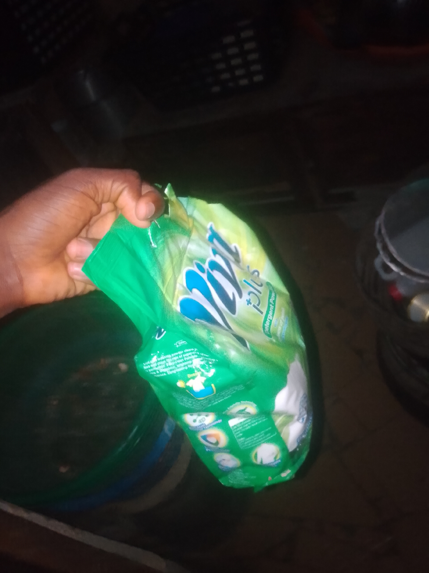 In Jauro Yinu, Nigeria, a person's hand is holding a green packet of detergent powder. The packet features white lettering and blue accents, with images of laundry depicted on its surface. The hand, with visible skin texture and fingernails, is positioned to the left of the packet. The background is dark and out of focus, with hints of a dark surface, possibly a bucket or container, to the left and a shadowy object resembling stacked items on the right. The lighting is dim, casting a shadow.