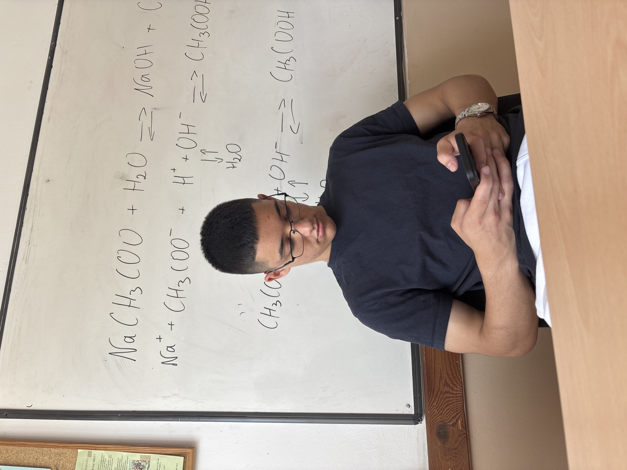 An adolescent male wearing a dark blue t-shirt and glasses is seated at a wooden desk in front of a whiteboard covered with chemical equations. He is looking down and interacting with a smartphone held in his hands, while a silver wristwatch is visible on his left wrist. The whiteboard displays equilibrium reactions involving sodium acetate, water, sodium hydroxide, hydrogen ions, hydroxide ions, and acetic acid. To the left of the whiteboard, a corkboard with papers is affixed to a white wall. The environment appears to be a classroom in Zlatograd, Bulgaria.