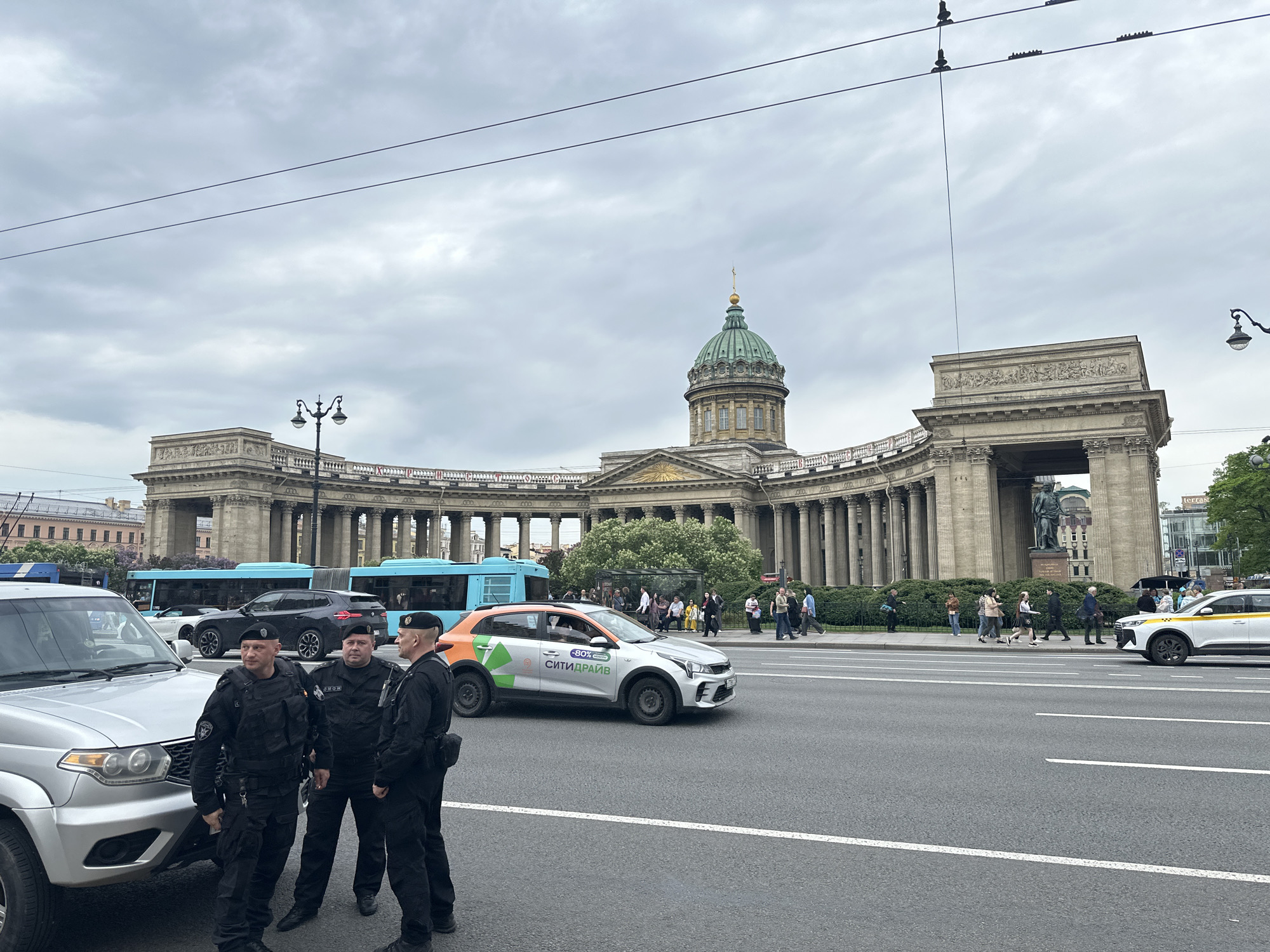 Three men in dark uniforms and berets stand at the edge of a road near a silver SUV. In the background is the Kazan Cathedral in Saint Petersburg, Russia. The sky is overcast. Traffic includes buses and cars. People are walking on the sidewalk in front of the cathedral.