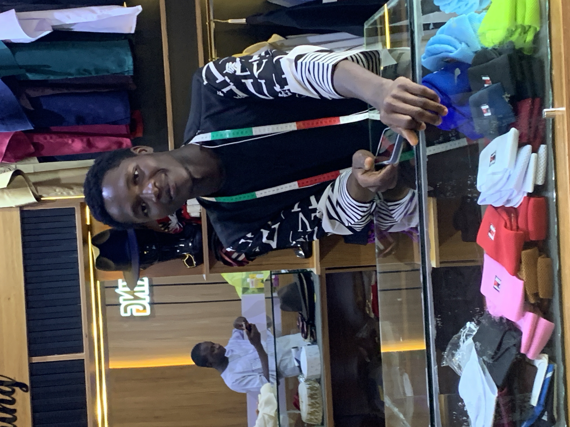 The image captures a young Black male, likely in his late teens or early twenties, standing behind a glass-fronted counter within what appears to be a clothing or tailor shop. He is wearing a black long-sleeved shirt with a white abstract pattern. A green, white, and red measuring tape is draped around his neck. He is holding a smartphone in his right hand, looking down at it.
The counter displays various folded items, including colorful caps (red, pink, blue, light blue, lime green), white cloths, and darker fabrics. Behind the counter, shelves are visible, with a mannequin wearing a dark hat on the upper shelf, and other clothing items hanging in the background. A sign with illuminated green text that reads "OMIE" is mounted on the wooden wall above the counter. Another person, a Black male dressed in a light blue traditional garment, is visible in the background on the left, also looking at a phone.
The environment suggests a commercial setting, likely a retail store specializing in apparel or custom tailoring. The lighting is warm and somewhat dim, with a prominent light strip illuminating the top shelf and a softer light source behind the "OMIE" sign. The overall impression is of a busy retail space, possibly located in Gura Top, Nigeria, given the context provided.
