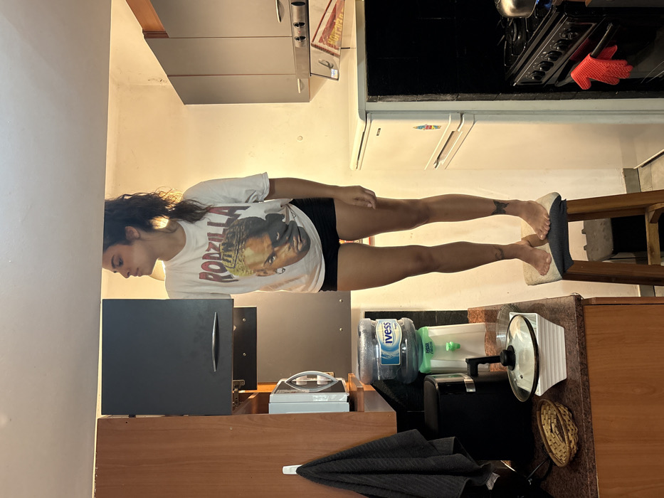A woman wearing a "Godzilla" t-shirt and shorts poses horizontally across a kitchen setting. Her feet rest on a stool. Above her are kitchen cabinets, a stove, and a refrigerator. Below her, a wooden counter holds a microwave, a water bottle, a blender, a coffee maker, and a dark umbrella. The image is slightly rotated counterclockwise.