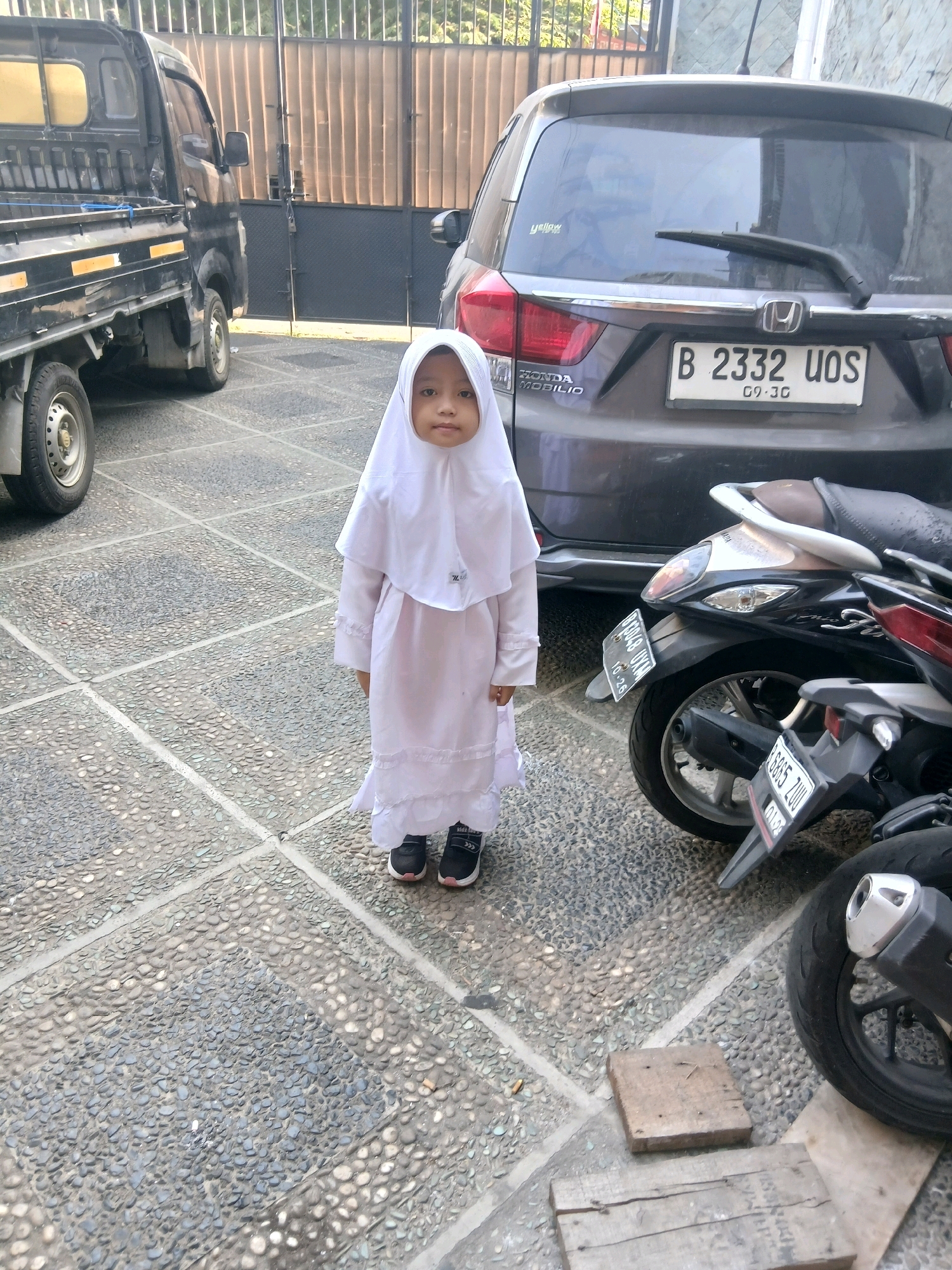 In West Jakarta, Indonesia, a young child wearing a white hijab and dress stands on a paved courtyard. The child is positioned between a black pickup truck and a dark grey car. Several motorcycles are parked behind the child, one of which is partially visible in the foreground. The courtyard is paved with light-colored cobblestones bordered by concrete. A metal fence and a building are visible in the background. The lighting suggests it is daytime.