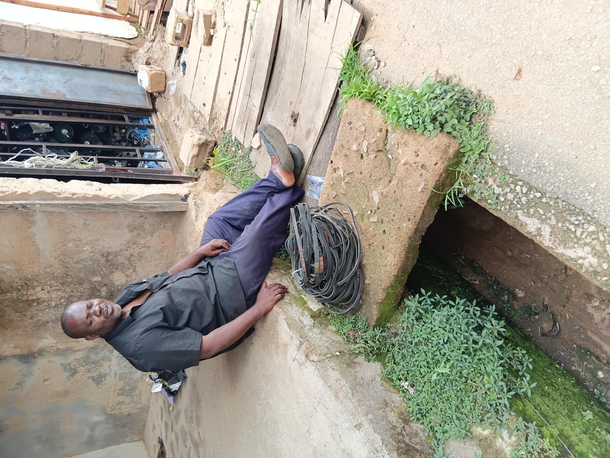 An adult male wearing a black short-sleeved collared shirt and dark trousers sits on a concrete ledge outdoors. He is positioned on the left side of the image, facing slightly to the right. His eyes are closed, and his mouth is slightly open. In front of him, a bundle of black belts is coiled on a stone slab that spans over an open drainage ditch. The ditch is partially overgrown with green vegetation. To the right of the man, wooden planks form a makeshift path. Behind him, a metal-grated door of a building is visible, with various items stored inside. The environment appears to be a street or alleyway in an urban setting, possibly in Jos, Nigeria, characterized by concrete structures, weathered walls, and evidence of everyday life.