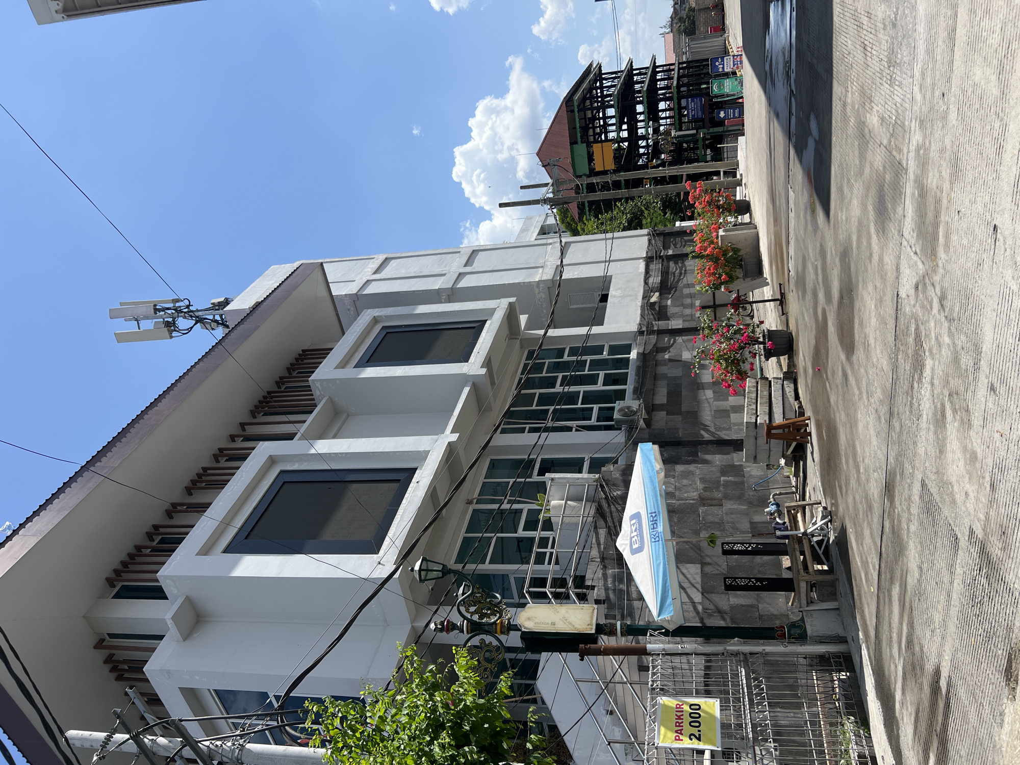 The image captures a street scene in the city of Yogyakarta, Indonesia, under a bright, clear blue sky with scattered white clouds. The foreground is dominated by a paved road with a textured, striped pattern, showing some wet patches. To the left, a modern, multi-story white building with dark-framed windows stands prominently. A green metal fence is visible in front of this building, with a yellow sign displaying "PARKIR 2.000" (Parking 2,000). Beside the fence, an ornate black lamppost with intricate scrollwork stands. An umbrella, emblazoned with the "BRI" logo, is open and positioned over a seating area with wooden benches and tables.
Behind the white building, to the right, is a dark, multi-level structure with a visible steel framework and a reddish-brown roof. This structure appears to be under construction or renovation. Further in the background, other buildings and trees are partially visible. The environment is urban, with visible electrical wires crisscrossing the scene. A few potted flowering plants with pink and orange blooms add a touch of color to the otherwise architectural landscape. No people are present in the frame. The overall impression is one of a quiet, sunlit day in a commercial or administrative area.