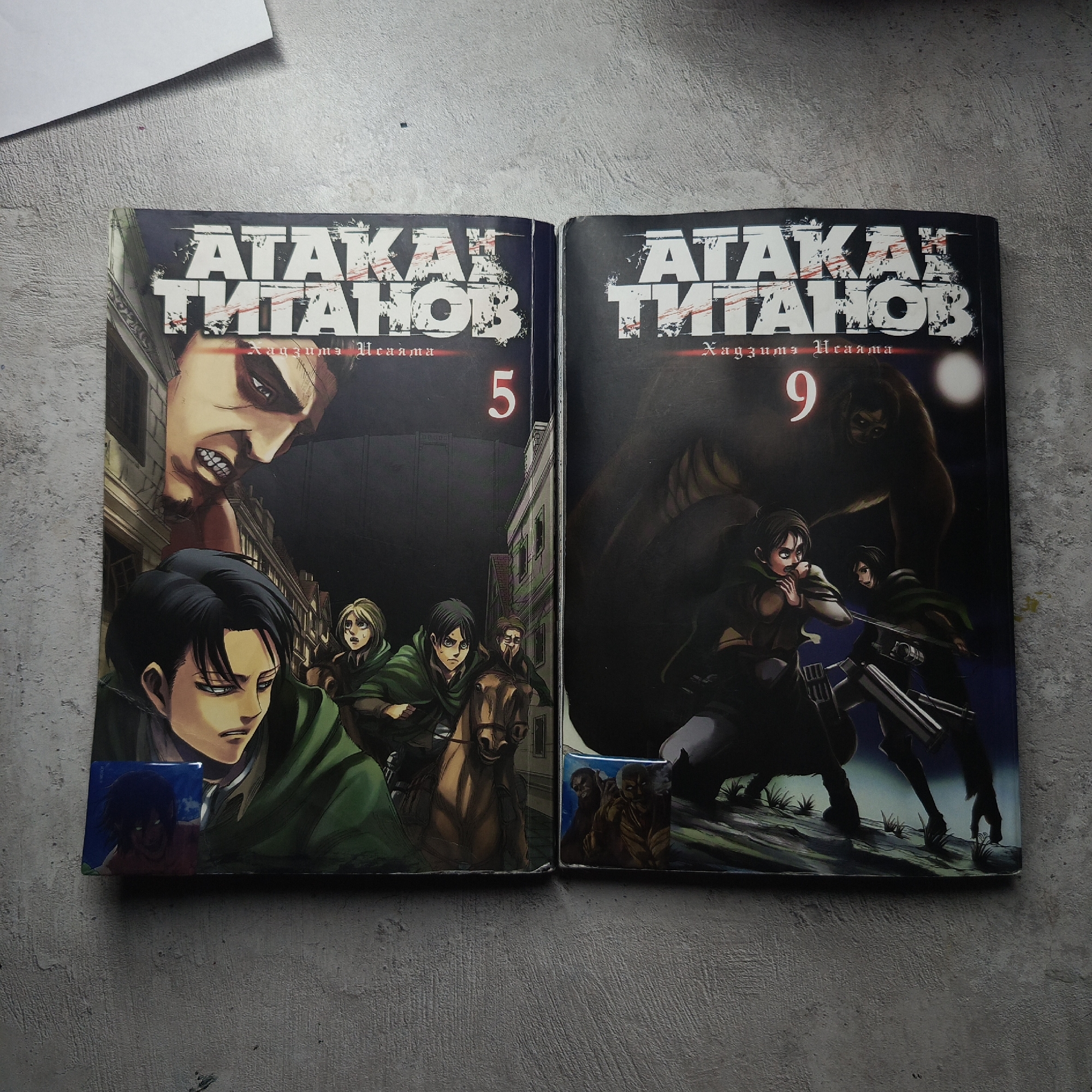 The image shows two manga volumes titled "Ataka Titanov" with volume numbers 5 and 9. Both are lying on a gray, textured surface. Volume 5 features a Titan face at the top, with characters including Levi and others in the lower section. Volume 9 depicts characters, including Eren and Mikasa, in action against a Titan at night. Both volumes have stickers adhered to their bottom corners.