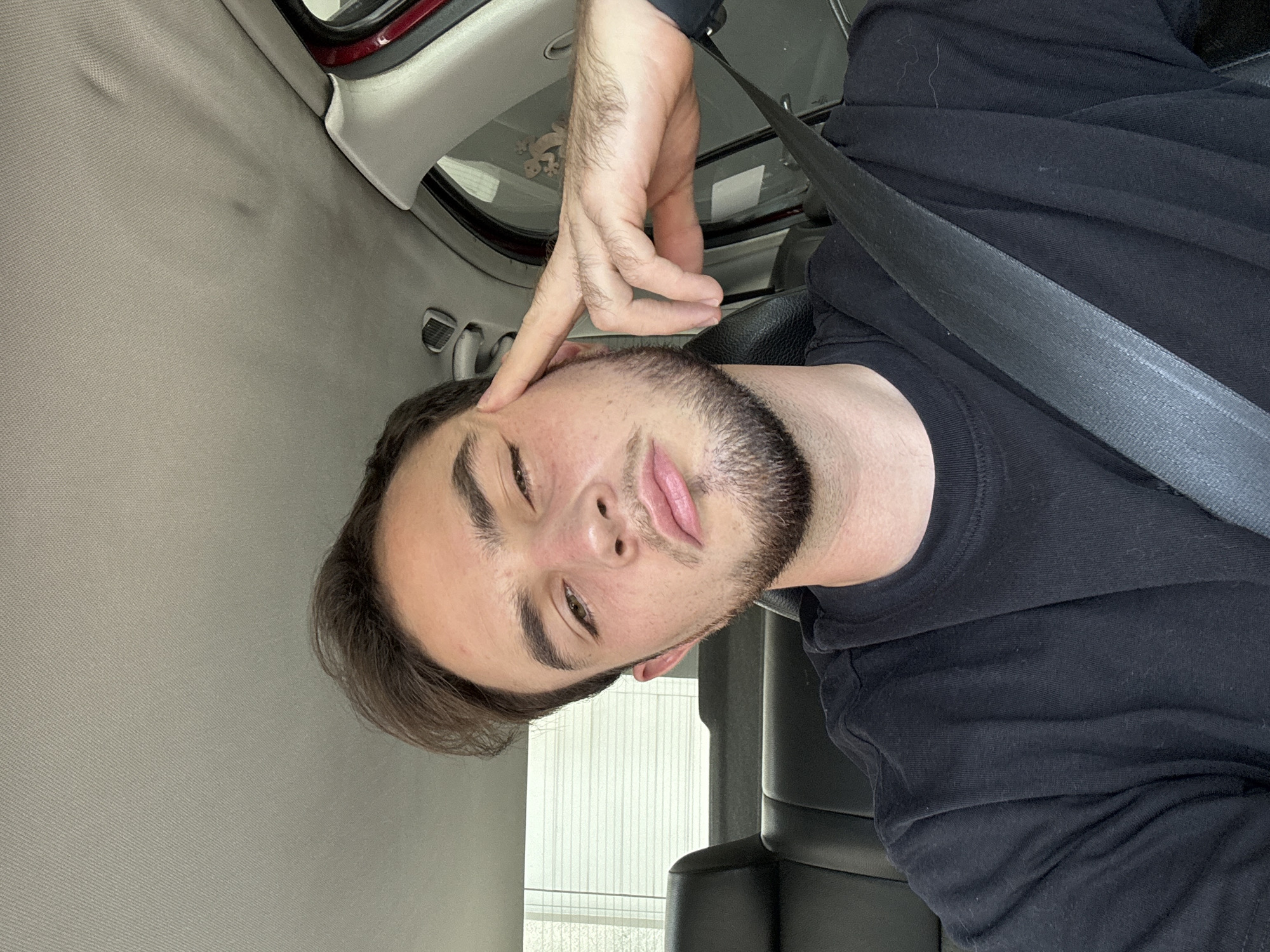 A selfie of a young man in a vehicle. He is wearing a black shirt and a seatbelt. His left hand is raised to his face, with fingers pointing at his forehead. The background shows the interior of a car, including the roof and a window. The location is likely inside a vehicle in San Bernardino Tlaxcalancingo, Mexico, but no specific elements confirm this.