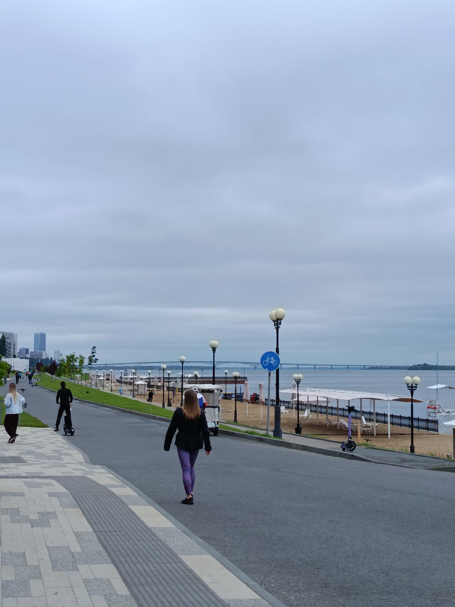 The image depicts a cloudy day at a waterfront promenade in an urban environment. In the foreground, a paved walkway with textured and smooth grey and tan tiles runs diagonally from the bottom left towards the center. A dark grey asphalt road is adjacent to the walkway, separating it from a sandy beach area.
Several people are visible. On the walkway, a woman with blonde hair wearing a light blue jacket and dark pants walks away from the camera. Further down the walkway, a person in a black outfit is riding a scooter-like device. On the road, a woman wearing a black jacket and purple patterned leggings walks away from the camera. Another person, dressed in black, is walking towards the camera on the road in the distance. On the beach, several figures are scattered among white beach chairs and shelters.
The beach area features white covered shelters and a dark fence. Along the edge of the road and separating the road from the beach, a line of lampposts with multiple globes is present. A blue circular sign with a white bicycle symbol is attached to one of the lampposts.
In the background, a body of water stretches towards a long bridge with visible supports. On the far side of the water, a tree-lined shore and the skyline of a city with several tall buildings are visible. The sky is overcast with grey clouds.