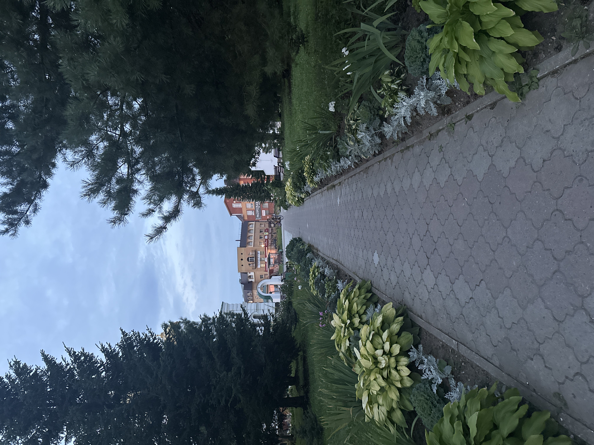 The image depicts a paved pathway leading into the distance, flanked by manicured flower beds and mature trees. The pathway is constructed of grey interlocking pavers with a decorative pattern. On either side of the pathway, there are garden beds filled with various plants, including large-leafed hostas with variegated yellow and green foliage, and silvery-grey leafy plants. Tall evergreen trees frame the scene, casting shadows. In the background, a cluster of multi-story buildings with pitched roofs is visible. One building appears to be light brown with red accents and has a sign that reads "РАСПРОДАЖ" (SALE). To the left of the pathway, a white building with a green arched entrance is partially visible. A person is seen riding a bicycle on the pathway in the distance. The sky is overcast with a blue-grey hue. The overall scene suggests a public park or walkway within a town or city. This location is identified as the town of Lysva, Russia.