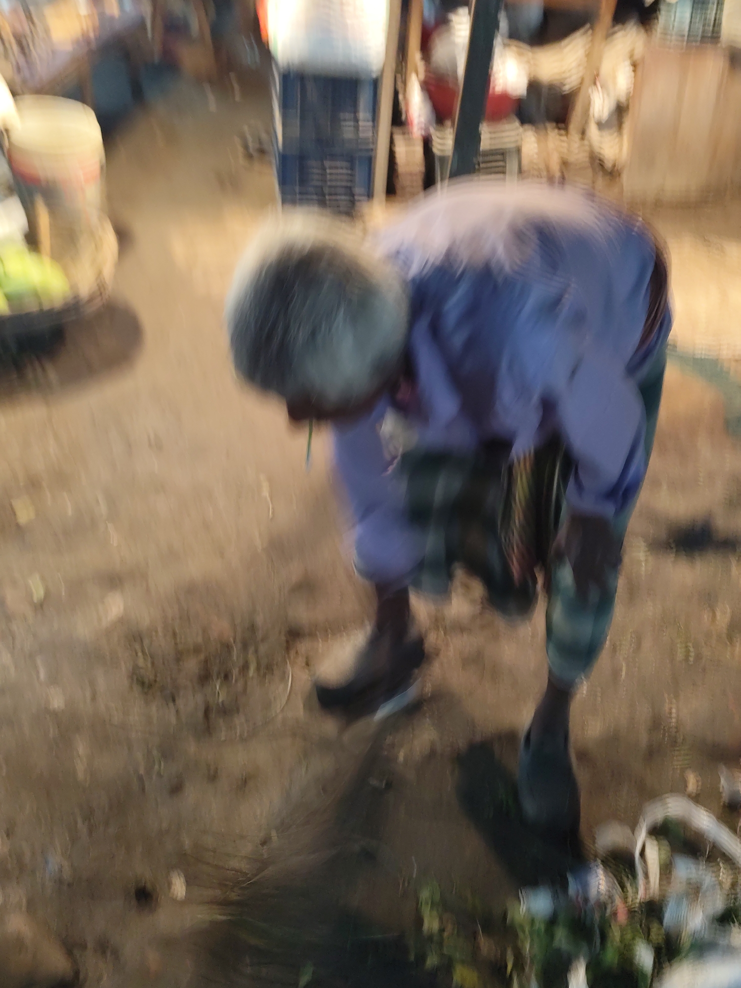 An elderly man with grey hair and a light blue long-sleeved shirt is bent over. He is wearing a checkered lungi. The scene appears to be outdoors on a dirt or concrete surface, with debris and scattered materials around him. A blurred object, possibly a cart with items, is visible to the left. The location is Dhaka, Bangladesh.