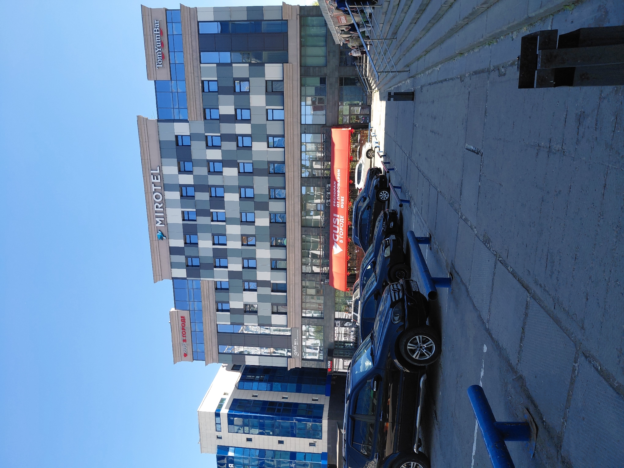 An outdoor, eye-level, wide shot shows a hotel in the city of Unknown, Unknown. The hotel is a multi-story building with a patterned facade of gray and white squares. The words "MIROTEL" are visible on the top of the building in large, white letters. To the right of "MIROTEL," the words "Tom Yum Bar" are also visible in white letters. Below the hotel, there is a red awning that reads "GUSI В ГОРОДЕ" in white lettering. In front of the hotel, there is a parking lot with several black SUVs and cars parked. The parking lot is made of concrete. To the right of the parking lot, there is a set of stairs leading up to a building. A few people are walking up the stairs. The sky is clear and blue.