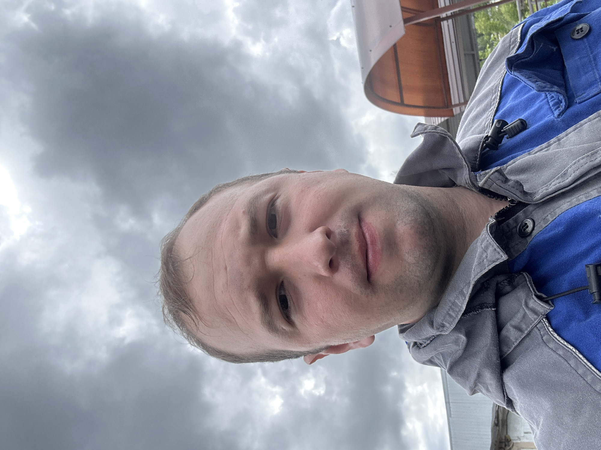 A close-up, portrait-style image features a man taking what appears to be a selfie outdoors. The man, with short light hair and stubble, looks directly at the camera with a neutral expression. He is dressed in a blue garment under a grey work jacket or overalls. A small black clip, resembling a microphone clip, is attached to the lapel area of his grey outer layer on his right side.
The background is predominantly an overcast, cloudy sky, indicating a dull or potentially rainy day. In the upper right portion of the frame, a translucent, light brown or orange arched structure, possibly a canopy, roof, or part of a small shelter, is partially visible. The lighting is soft and diffused due to the heavy cloud cover. There is no visible text. The setting is an outdoor location in Chaikovsky, Russia.