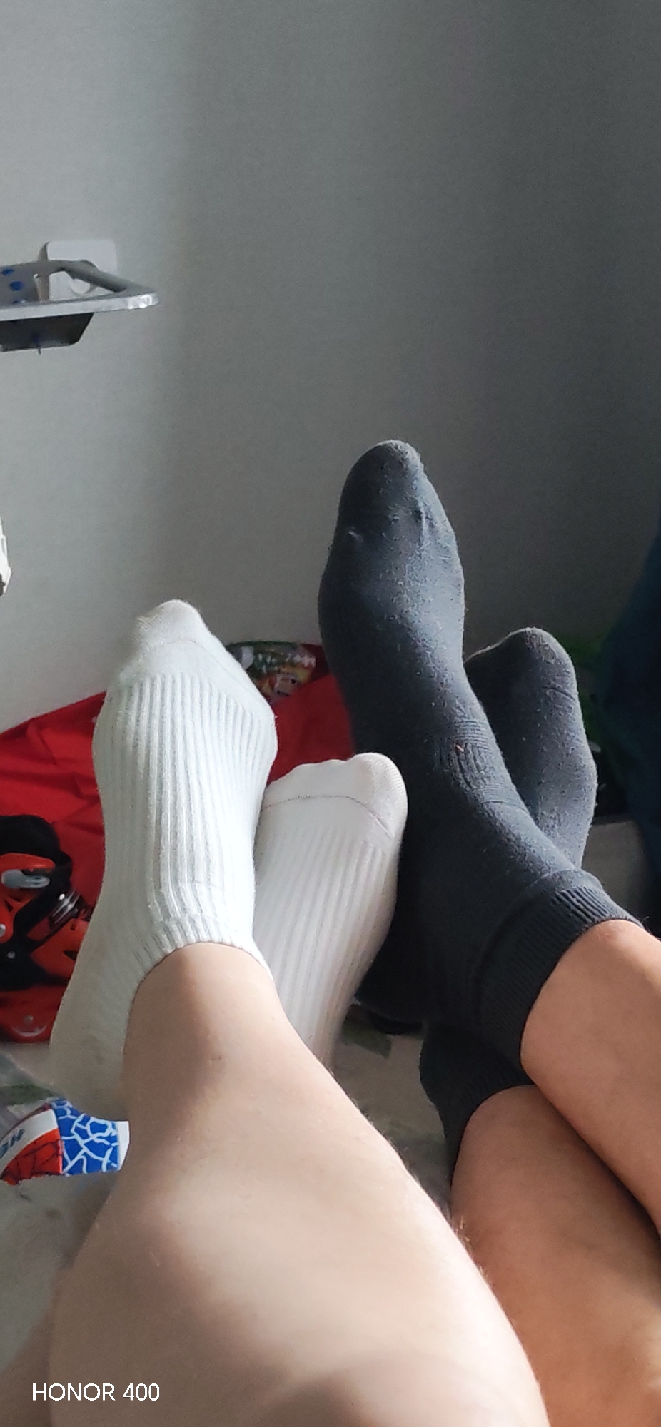 Two pairs of feet, one wearing white ribbed socks and the other wearing gray socks, are angled towards the camera on what appears to be a bed. A red object with a mask-like graphic, possibly a pillow or blanket, is partially visible behind the white-socked feet. A blue and white object is visible on the lower left. A wall is partially visible in the background. "HONOR 400" is printed at the bottom left. I have no information about the location.