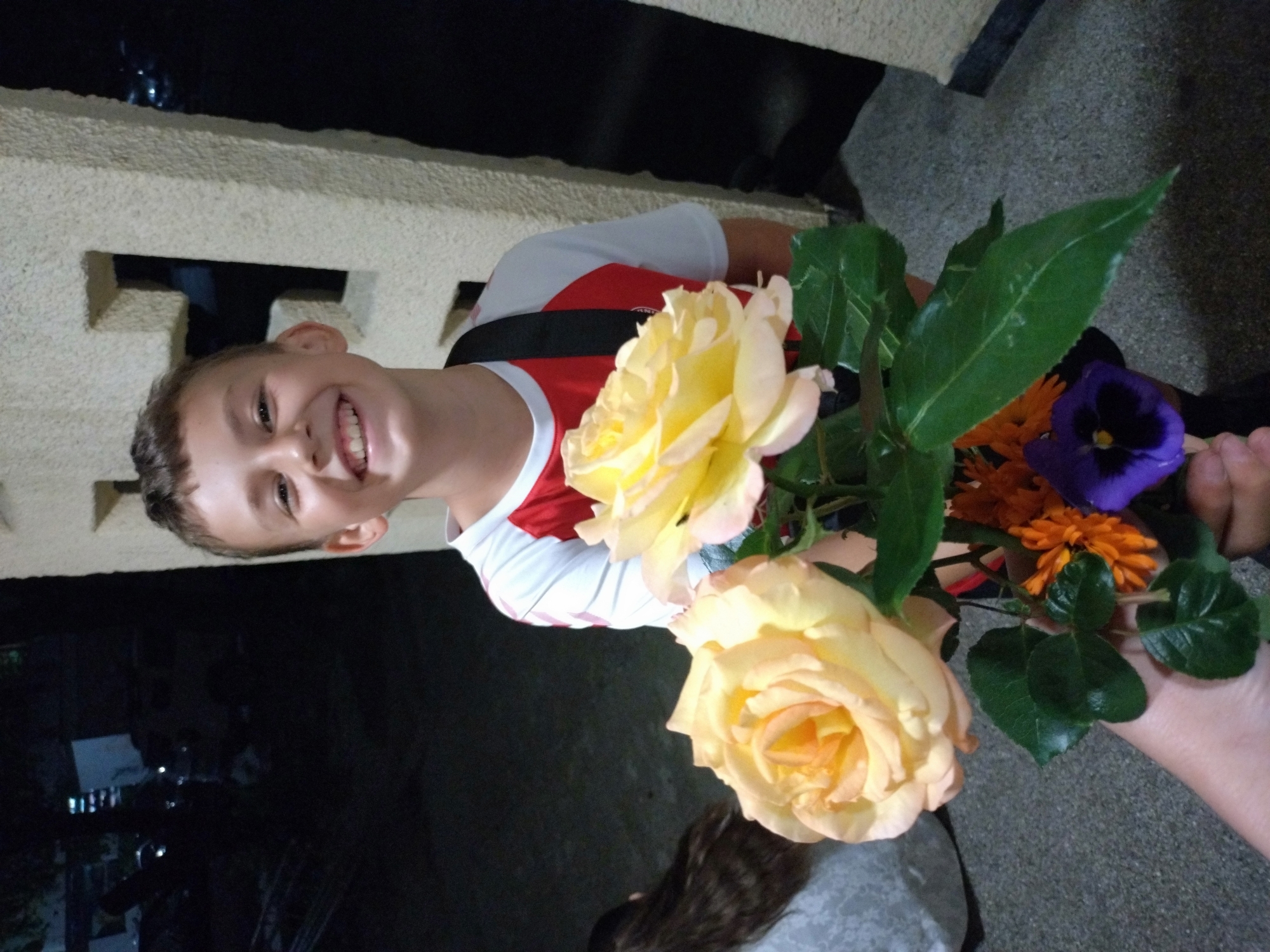 A young boy holds a bouquet of flowers in front of a stone building. The boy is smiling and wearing a white shirt with red sleeves and a black strap. The bouquet contains two large yellow roses, orange flowers, and a purple flower. The background shows a building with a stone wall and an opening. The location is in the city of Stara Zagora, Bulgaria.