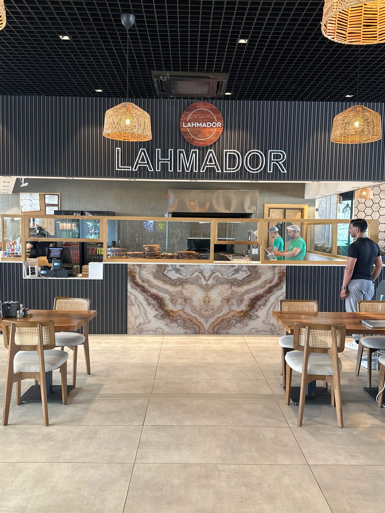 The image depicts the interior of a restaurant called LAHMADOR, located in Guzelyurt, Northern Cyprus. The restaurant features a modern design with a black slatted wall as a backdrop. A large, prominent sign with the restaurant's name is displayed above the counter area. Woven pendant lights hang from the black tiled ceiling, casting a warm glow.
In the foreground, there are several dining tables and chairs. The tables are made of wood with black metal legs, and the chairs have wooden frames with woven rattan backs and grey upholstered seats. One table has a laptop on it.
The counter area showcases a marble-patterned wall that serves as the backsplash for the service counter. Behind a glass partition, two restaurant employees wearing green shirts and hairnets are visible working. The counter itself is equipped with a point-of-sale system, cash register, and various food service items. A man in a black t-shirt and grey pants stands facing the counter area, looking towards the workers.
The floor is tiled with large, light-colored square tiles. The overall impression is of a clean, contemporary dining establishment.