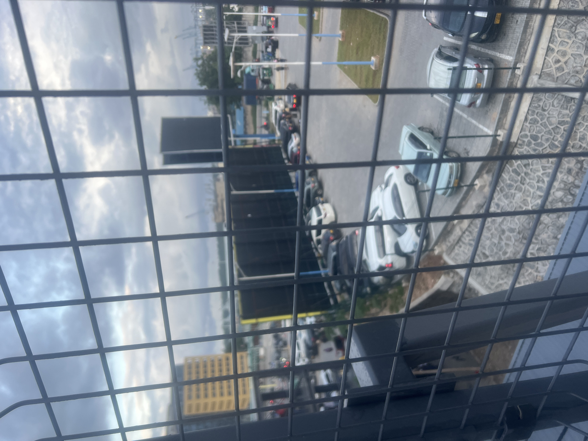 The image, viewed through a metal grate, shows an urban landscape. Cars are parked in a lot next to a paved area. Beyond the parking lot is a green space with blue posts. Further in the background, industrial structures and trees are visible under a cloudy sky. A tall, yellow building can be seen in the lower left. The location is likely Dar es-Salaam, Tanzania.
