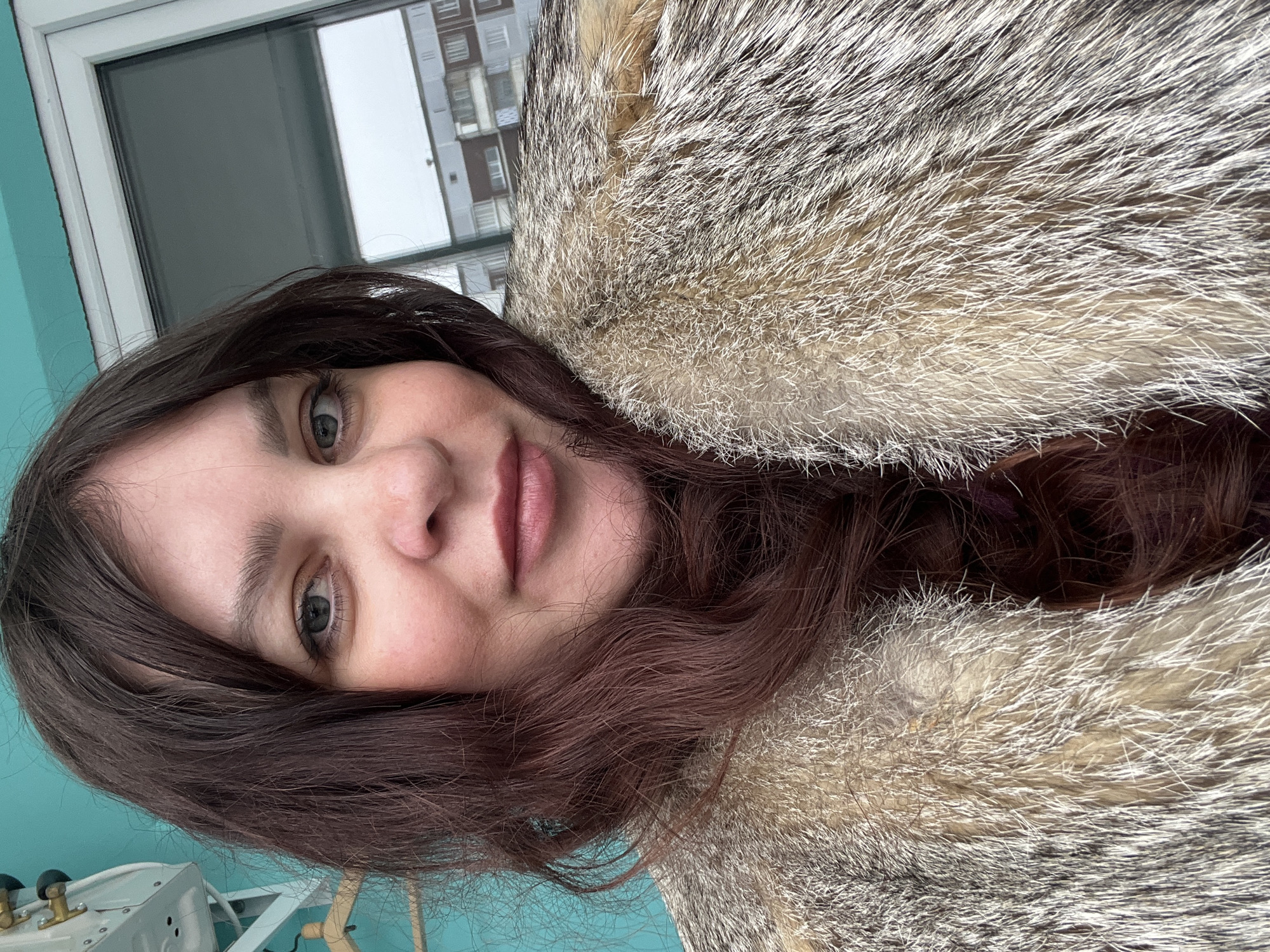 A selfie shows a woman wearing a fur coat, with the city of Perm, Russia, visible through a window in the background. The woman has brown hair and light skin, and she is looking at the camera with a neutral expression. The fur coat is gray and white, and it appears to be quite warm. The background shows a building with many windows. It is likely that this photo was taken in winter, given the presence of the fur coat and the weather conditions outside.