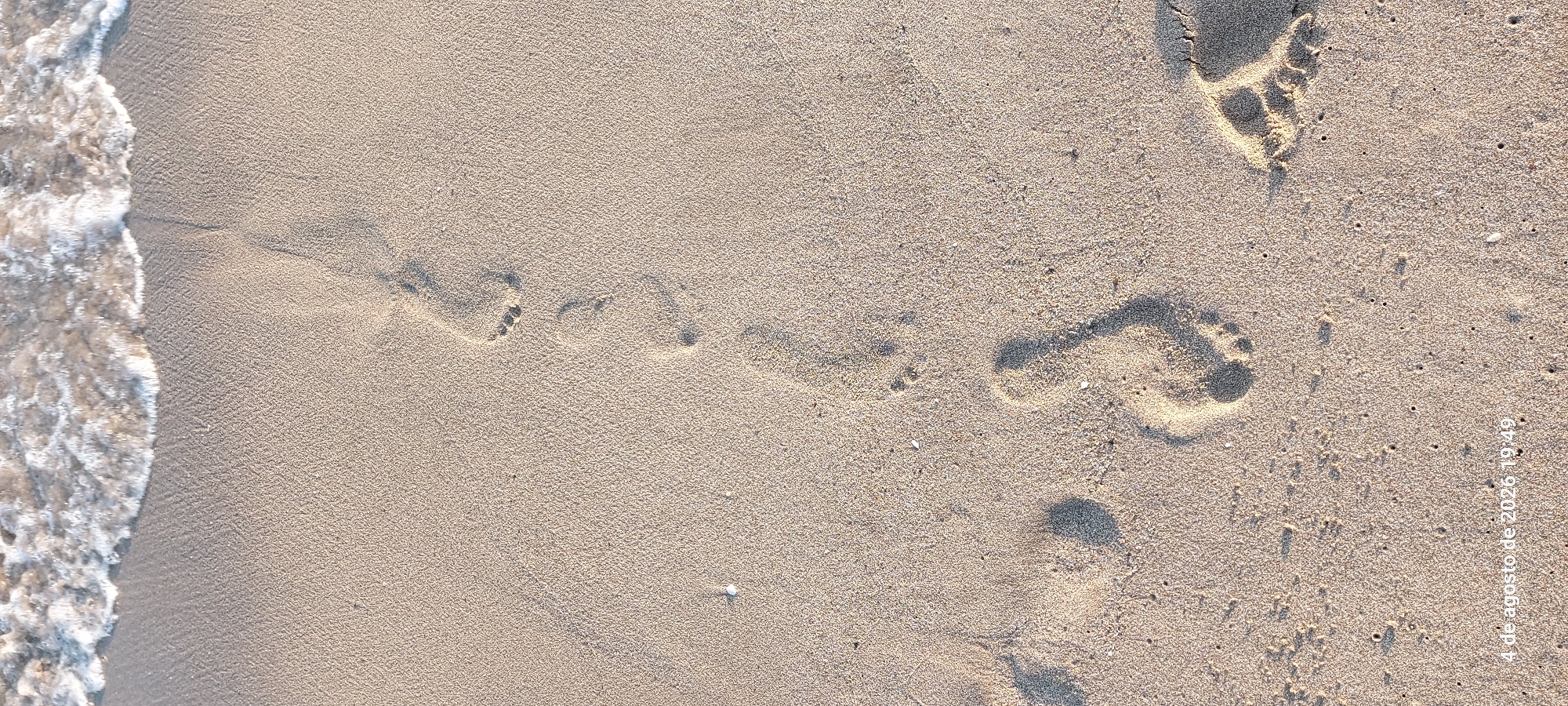 The image shows a sandy beach with footprints leading away from the gentle lapping waves. The sand is a light beige color and appears damp near the water's edge. The footprints are clearly defined, indicating recent passage. The overall scene suggests a tranquil beach environment. The timestamp "4 de agosto de 2026 19:49" is visible in the lower left corner, indicating the image was taken in Castelldefels, Spain.