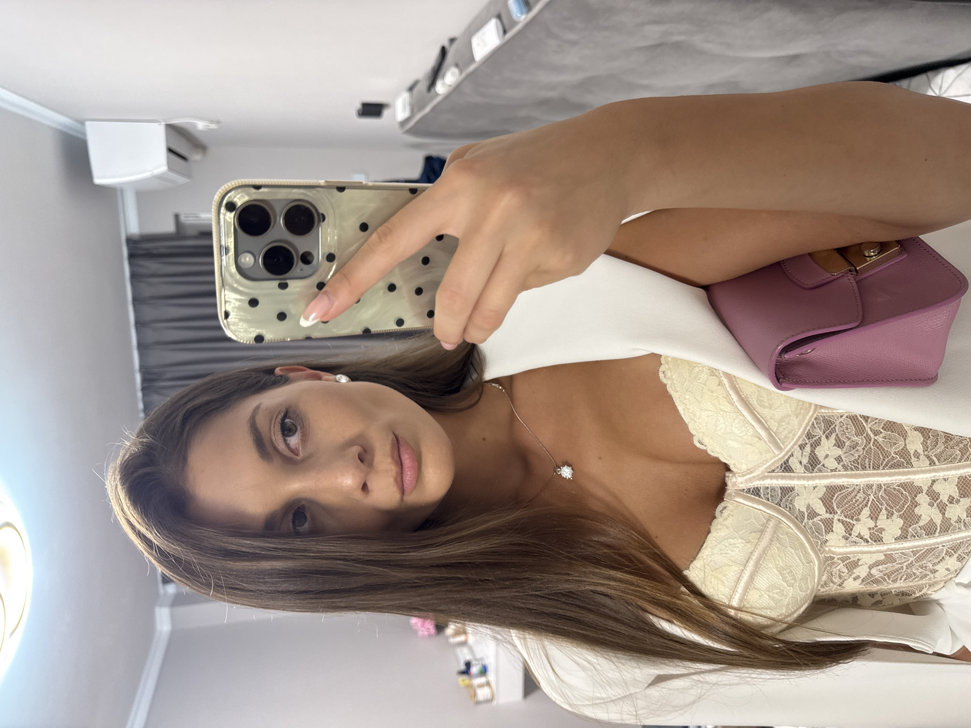 A woman is taking a selfie in a room with white walls. She is holding an iPhone with a black polka dot case. She has long brown hair and is wearing a white top with lace and a white jacket. She is also wearing a silver necklace with a diamond pendant. A lavender purse is visible near her. In the background, there is an air conditioner and a grey headboard of a bed. The location is likely indoors, but there is no indication of being in Burgas, Bulgaria.