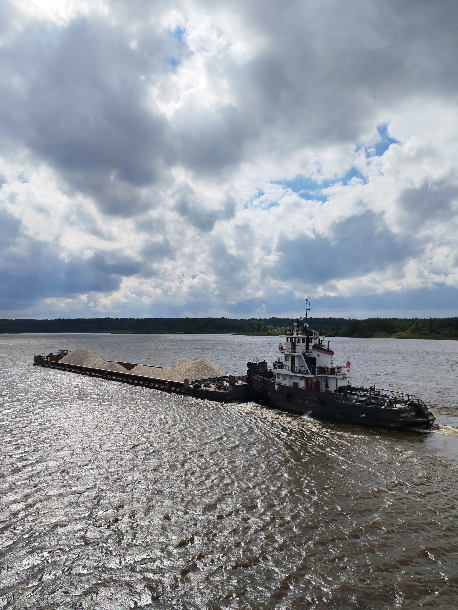 An aerial, eye-level, wide shot shows a tugboat pushing a barge laden with gravel down a wide, dark river. The barge is to the left of the tugboat. The water is choppy and reflective, with sunlight glinting off the waves. The sky is mostly filled with dark, heavy clouds, with a few patches of blue visible. A dark, tree-lined bank of land is visible in the distance. The tugboat is dark-colored with a white superstructure. It has a red stripe along its side and a flag flying from its mast. The barge is long and flat, filled with light-colored gravel. The tugboat is moving away from the camera, to the right. The scene is serene and industrial, suggesting transport and labor.