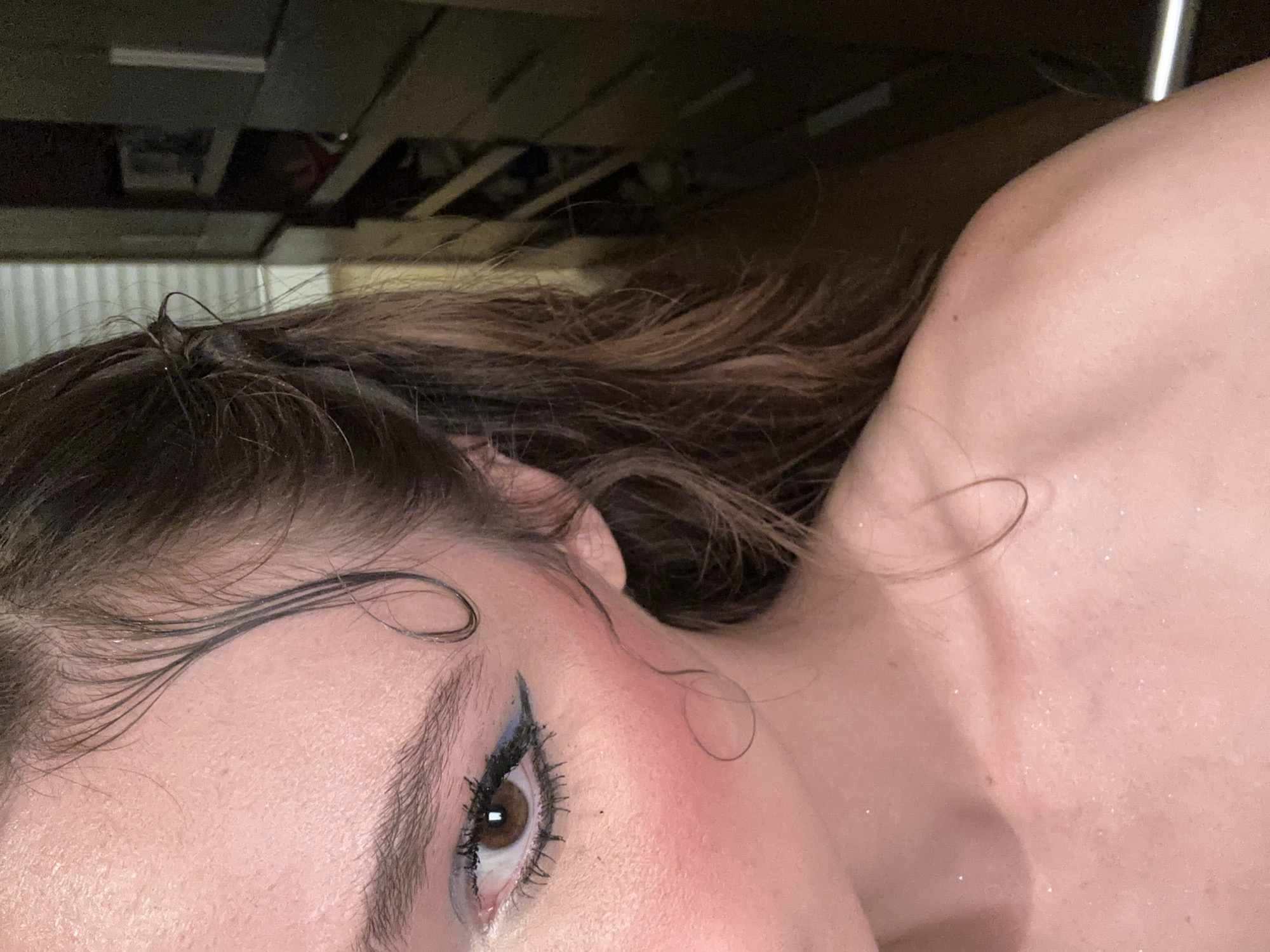 The image shows a close-up, upper-body, side-profile of a young woman. She has brown hair styled with wet tendrils framing her face and swept back. Her eye is visible, with dark lashes, brown iris, and blue eyeshadow with black winged eyeliner. Red blush is applied to her cheek. Her shoulder and upper chest are also visible, with faint glitter on her skin. The background is blurred, showing what appears to be a dresser or closet with shelves and drawers. The lighting suggests an indoor setting. There is no discernible indication of the location being in San Bernardino Tlaxcalancingo, Mexico.