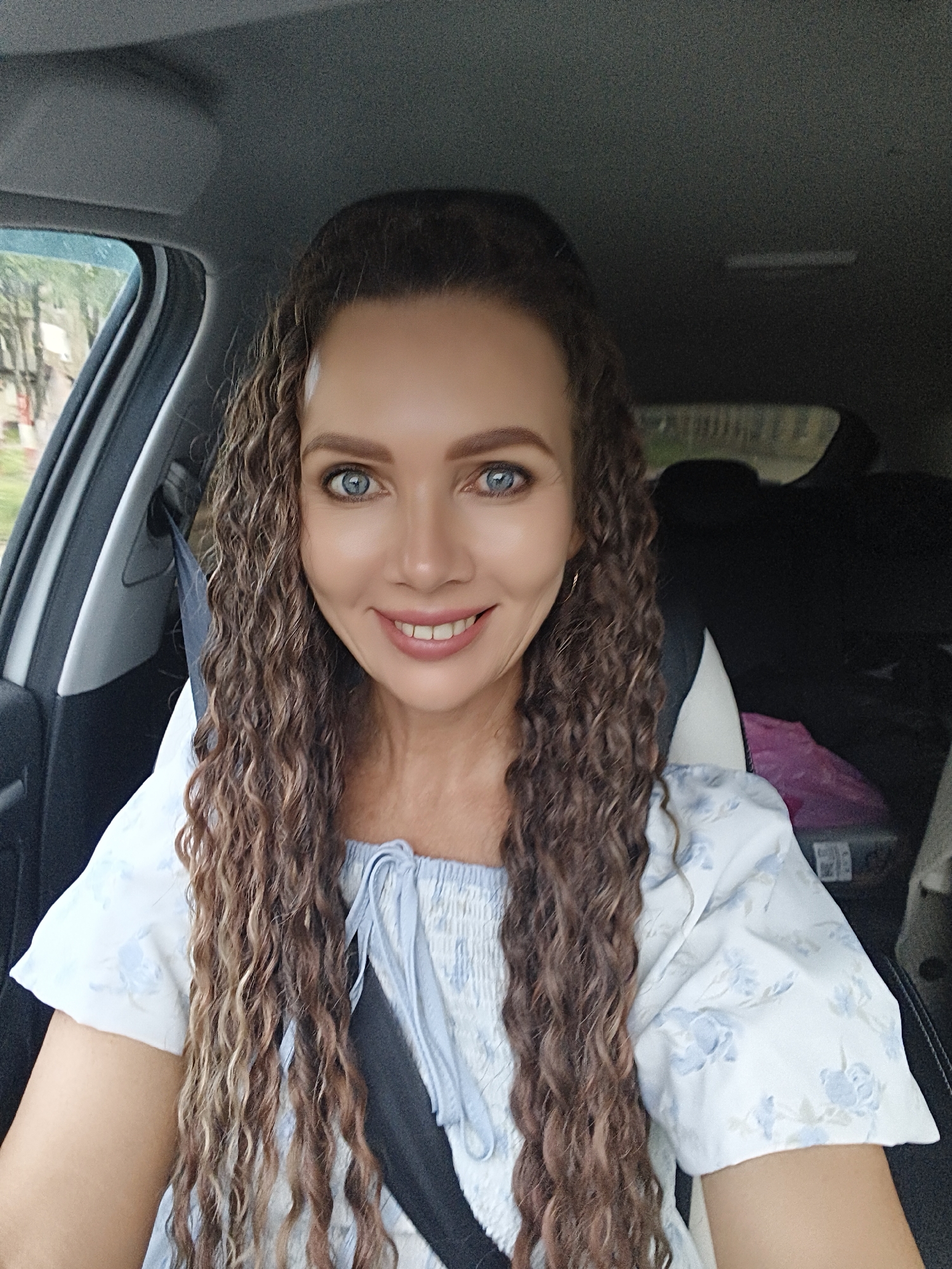 A woman with long, curly brown hair and blue eyes is sitting in the driver's seat of a car. She is smiling at the camera and wearing a white blouse with a blue floral pattern. The car's interior is visible, including the dashboard and a seatbelt. The background outside the car windows suggests a natural, possibly rural setting with trees and buildings. The image appears to have been taken during the daytime.