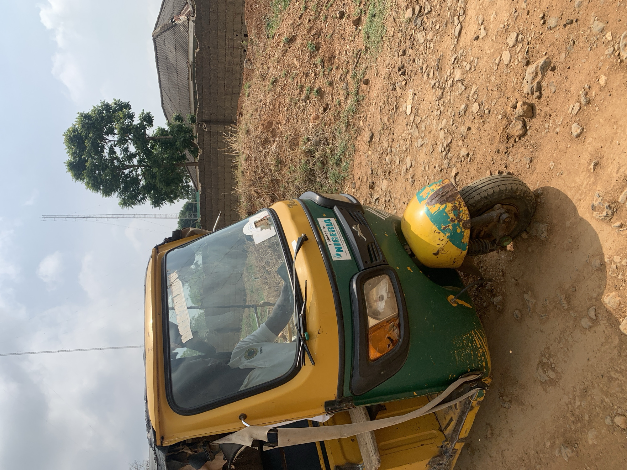 The image displays a yellow and green three-wheeled auto-rickshaw, commonly referred to as a Keke Napep, parked on a dry, unpaved dirt path. The vehicle is oriented slightly towards the right, with its front wheel and a somewhat worn, yellow fender, showing chipped paint, in the foreground. The main body of the vehicle is green and yellow. On the green section above the windshield, a sticker is visible with "NIGERIA" and the partially legible phrase "I pledge to". Inside the vehicle, a person can be glimpsed wearing a white shirt that features a logo, though the text on it is unclear. Makeshift strapping or repair materials are visible near the lower front chassis of the vehicle.
The immediate surroundings consist of reddish-brown, dusty earth, interspersed with numerous small rocks and sparse patches of dry grass. In the background, a large, verdant tree stands prominently against a bright, clear sky. To the right of the tree, part of a building constructed from concrete blocks with a dark, corrugated metal roof is visible. Further in the distance, a tall, slender antenna or communication mast can be seen. The bright sunlight suggests it is daytime, under clear weather conditions. The scene depicts a stationary vehicle in what appears to be a rural or semi-urban setting, consistent with the provided location context of Maigizo, Nigeria. No specific activity or event is taking place beyond the vehicle being present on the path.