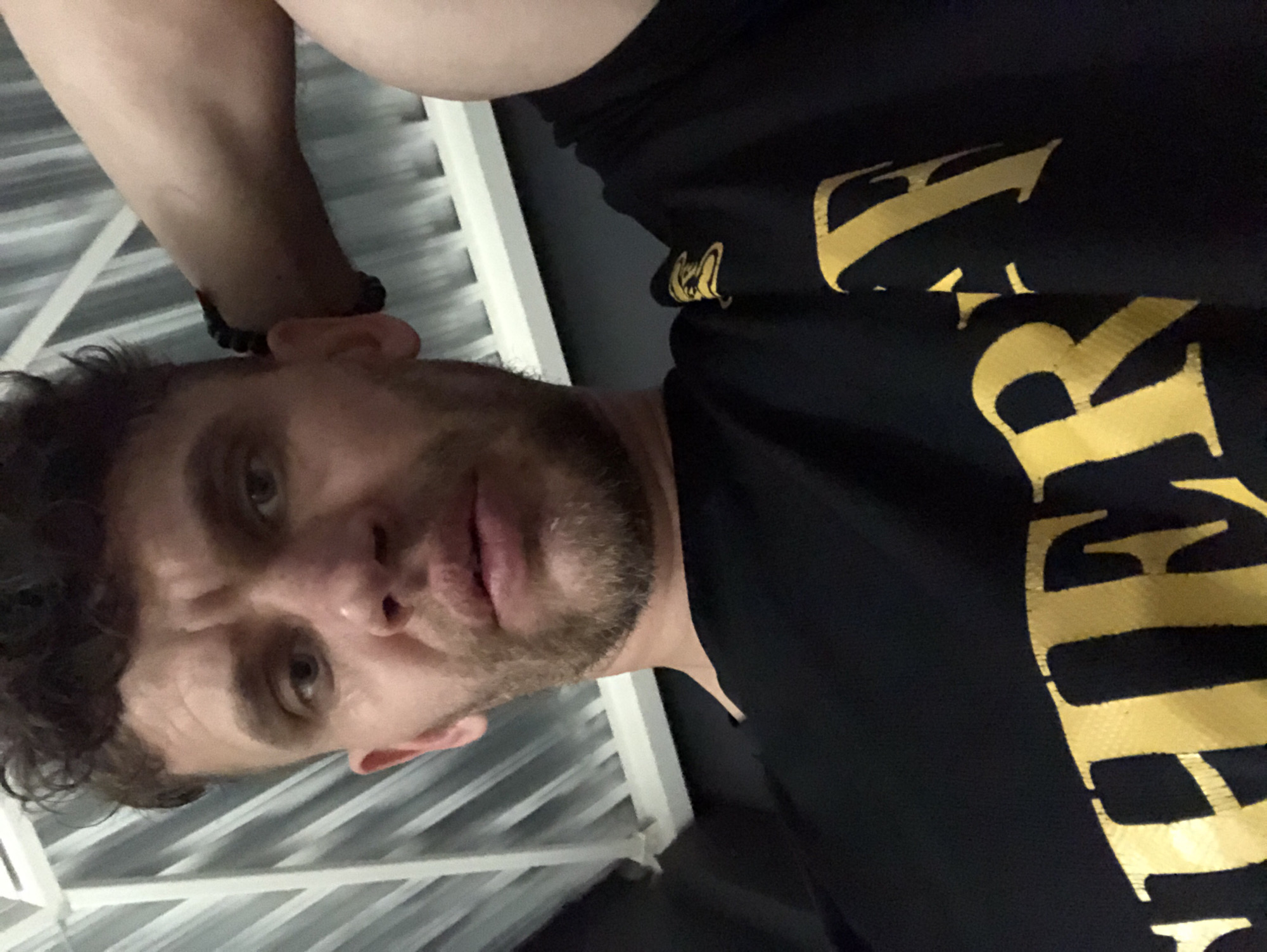 A man is captured in a low-angle, close-up selfie. He has short, curly, dark brown hair, a light complexion, and a short beard. His eyes are open and looking towards the camera. He is wearing a black t-shirt with a yellow graphic. His right arm is bent, with his hand resting behind his head. The background appears to be an indoor setting with a corrugated metal ceiling or wall. The lighting is dim, with a light source above and to the left. There is no identifiable information suggesting a specific location in Saint Petersburg, Russia.