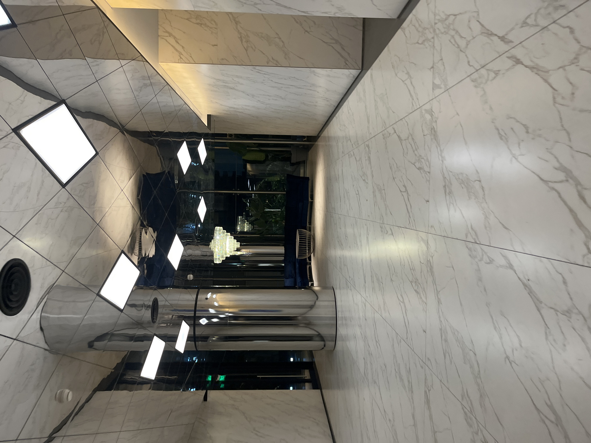The image depicts an interior space with marble-tiled walls and floors. A cylindrical metallic structure stands in the foreground, reflecting the surroundings. Rectangular light fixtures are integrated into the ceiling and walls, providing illumination. In the background, a glimpse of greenery suggests an outdoor or garden area visible through a glass partition. A chandelier is visible in the distance, indicating a luxurious or decorative element within the space. The image does not contain people, and there are no identifiable actions or interactions taking place. The scene appears to be a modern, upscale lobby or hallway. The image does not provide enough details to infer the exact location in Buenos Aires, Argentina.