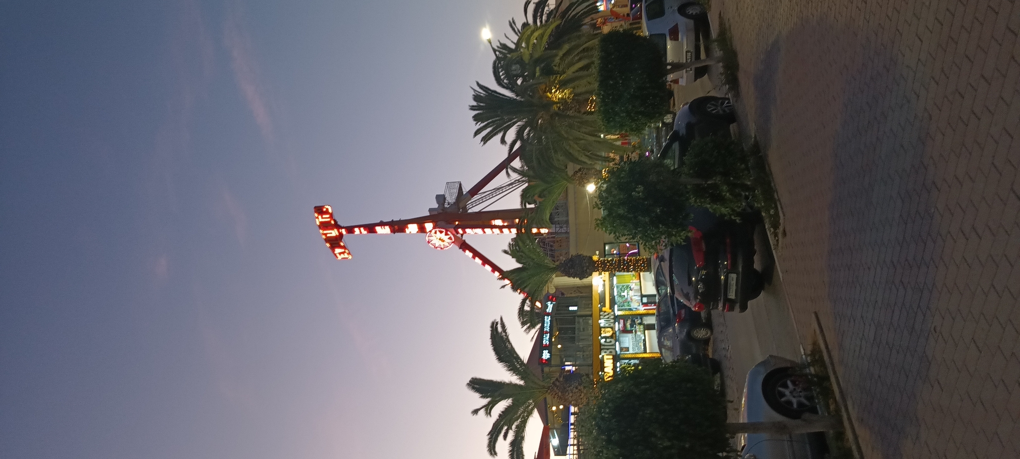 This is a vertical, full-shot photograph taken at dusk in Hammam Sousse, Tunisia, depicting an amusement park setting. In the background, a tall amusement ride with red and white lights is prominently displayed against a gradient twilight sky. The word "TITUS" is illuminated in red letters at the top of the ride. Palm trees frame the ride and buildings below, suggesting a warm climate. In the middle ground, a building with yellow signage reads "Restaurant BIG BOSS" and "SPACE AMOUR". Several cars are parked in front of the restaurant. In the foreground, meticulously pruned green bushes and trees line a paved walkway. The overall impression is one of a lively entertainment district at the close of day.