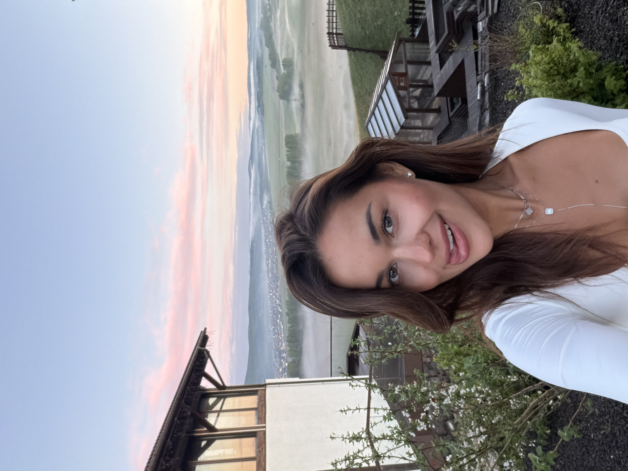 A smiling woman with brown hair, wearing a white long-sleeved shirt and a delicate necklace, poses for a selfie. The background features a misty valley at dawn or dusk, with building roofs and trees visible. The location is likely near a building with a wooden structure and possibly a greenhouse-like extension. The scene does not appear to be in Russia but could be in an area with similar landscapes. "Shamoni" appears to be an incorrect city name.