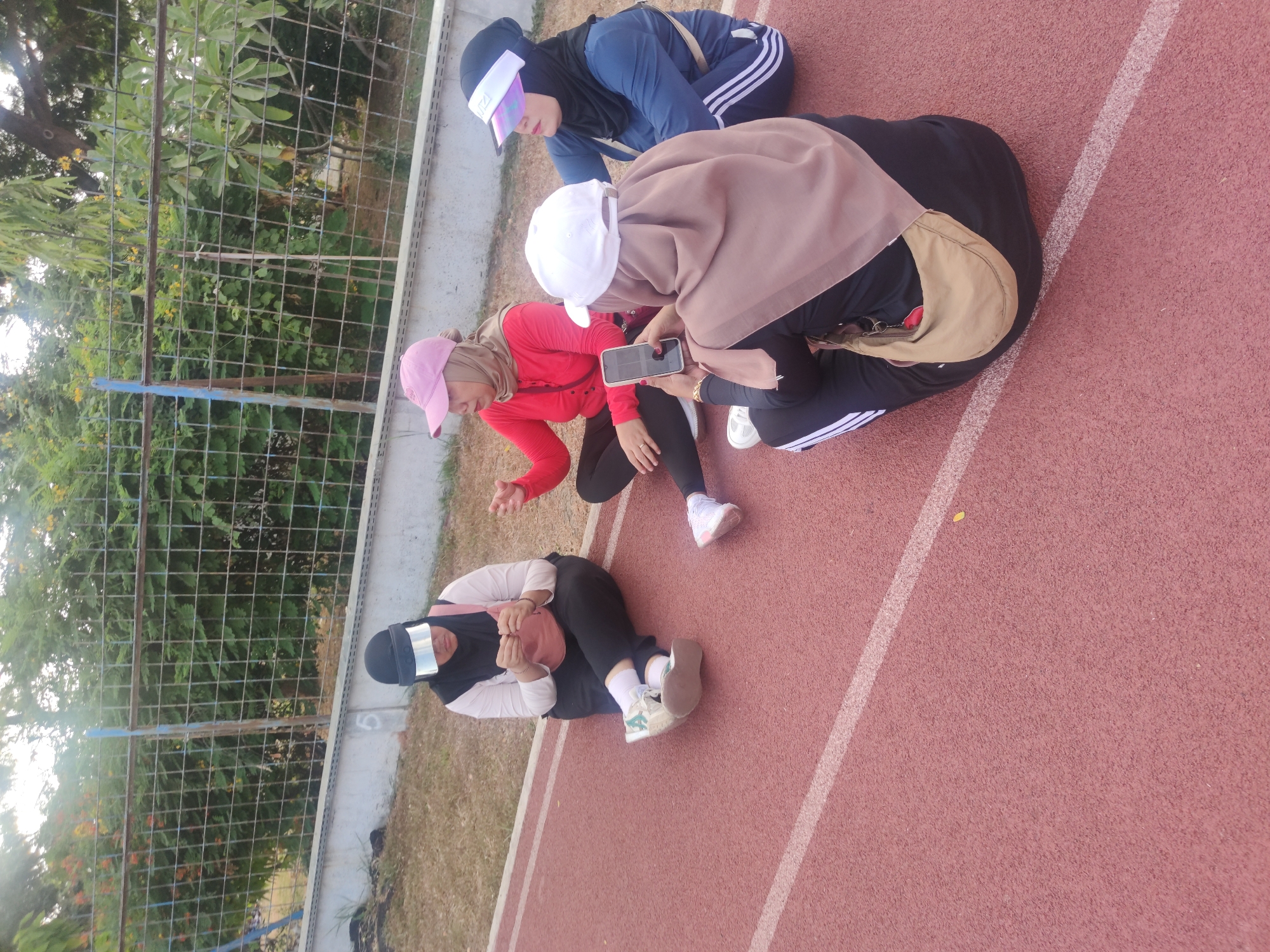 Four individuals, appearing to be women, are seated on a red running track, possibly in a park or sports facility within Surabaya, Indonesia. The individuals are wearing athletic attire, including headscarves and visors. One person is holding a mobile phone, and the others are engaged in conversation. The background features a metal fence and lush greenery, suggesting an outdoor setting. The lighting indicates it is daytime, with sunlight filtering through the trees. The running track itself is clearly visible, marked with white lines.