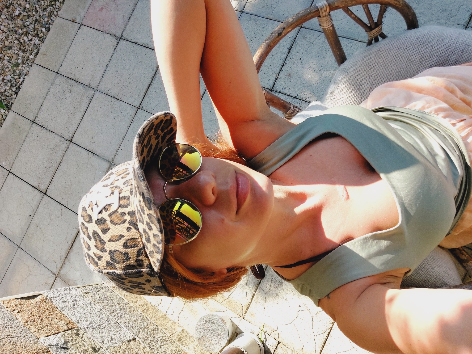 In Sozopol, Bulgaria, a woman with auburn hair and a leopard print baseball cap and round sunglasses is captured in a selfie. She wears a light olive-green tank top and a pale peach skirt. The woman is seated on a wicker chair with a woven back, partially covered by a gray blanket, outdoors on a paved patio. Sunlight casts strong shadows across the scene. A stone wall is visible on the left, and gravel is scattered on the ground in the upper right quadrant.
