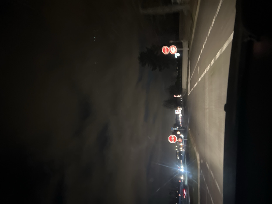 A nighttime view from a car driving on a road with clear lane markings. Several illuminated road signs, including stop signs, are visible along the side of the road. Other cars' headlights pierce through the dark background. The sky appears cloudy or foggy. The scene conveys a sense of motion and darkness.