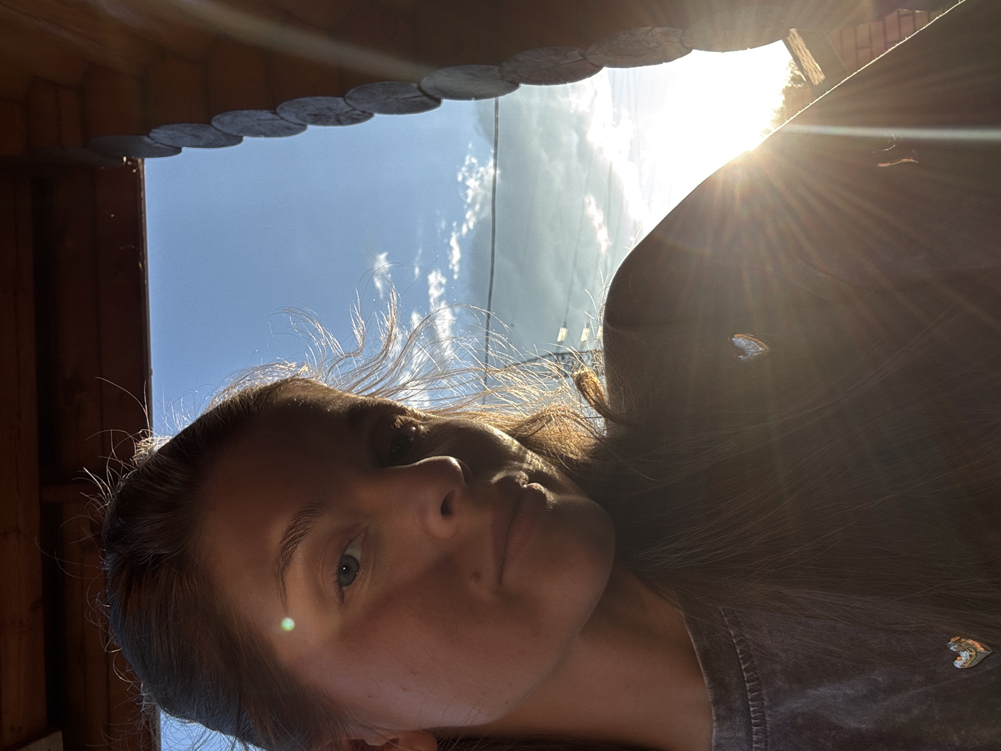 This media file is a portrait of a young woman looking towards the camera. She has fair skin, light blue eyes, and long, dark brown hair that is slightly windblown. The sunlight is casting a warm glow on her face, creating lens flares and highlighting her features. She is wearing a dark, textured shirt with a small, heart-shaped embellishment on the left side.
The setting appears to be outdoors, possibly on a porch or under a wooden structure, indicated by the wooden planks visible at the top of the frame. The background is dominated by a bright, blue sky with some scattered clouds, suggesting it's a clear and sunny day. The sun is intensely bright, positioned high in the sky and to the right of the frame, creating a halo effect and strong rays of light. This indicates it is likely midday or early afternoon.
The overall mood is serene and possibly contemplative, with the natural lighting contributing to a peaceful atmosphere. There is no visible text in the media file. The context provided places this scene in Minusinsk, Russia.