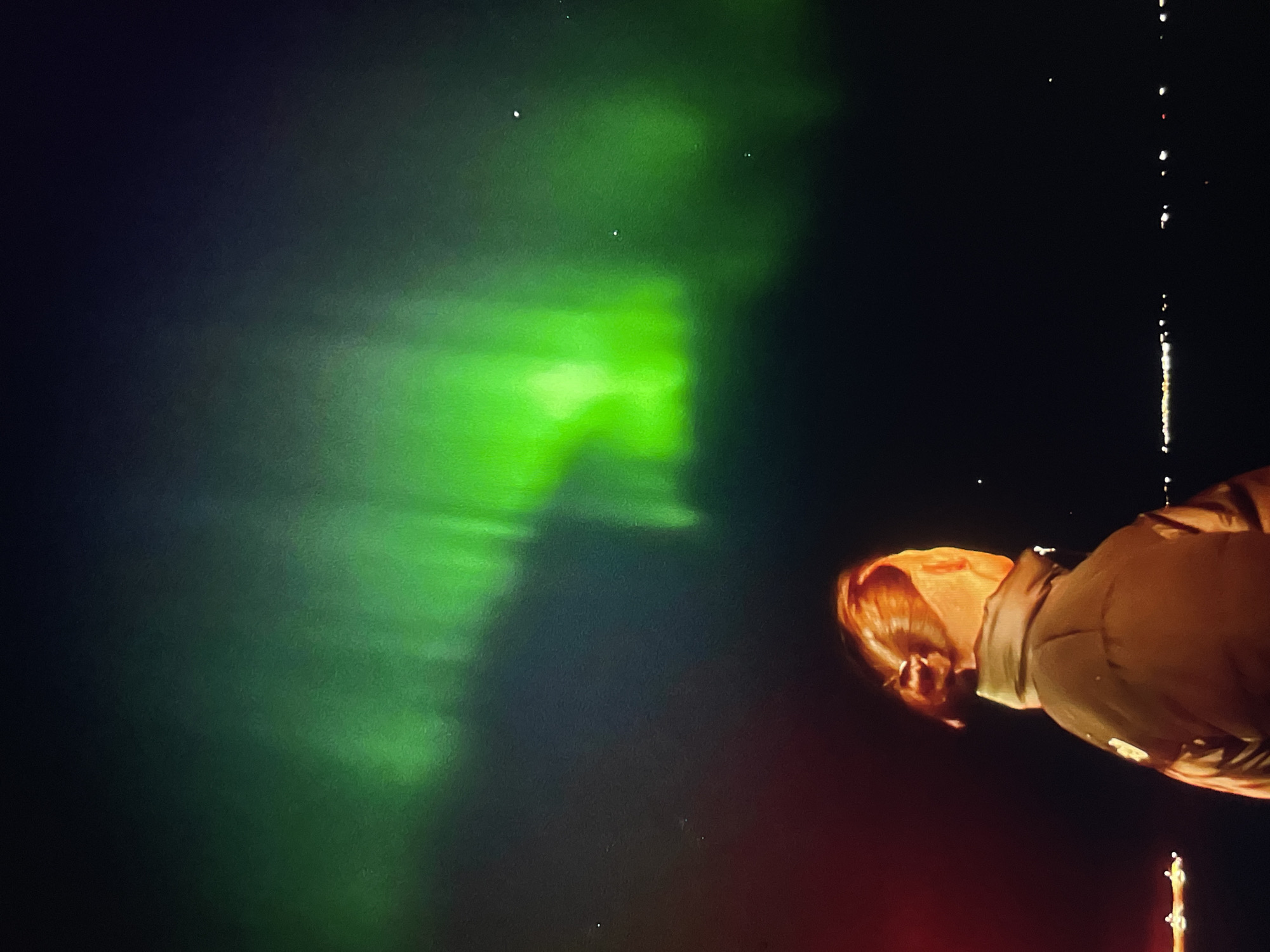 The image shows a person standing at night under the aurora borealis. The aurora appears as a bright green band across the upper left portion of the frame. The person is wearing a jacket. The location is Unknown, Unknown.