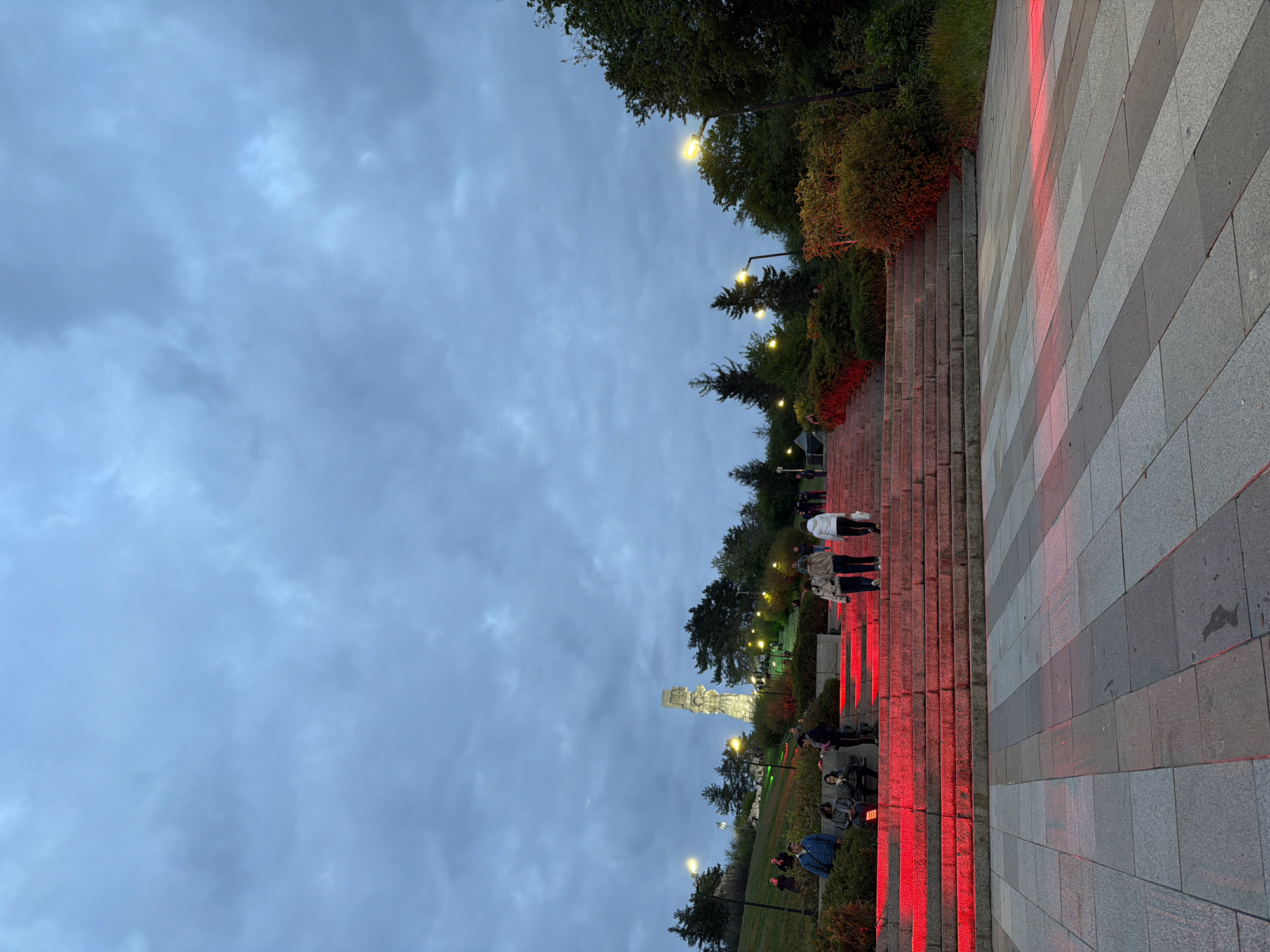 An evening scene captures an outdoor public space, likely a memorial park or monument complex. A grand staircase, illuminated with striking red lights along its edges, dominates the right side of the frame, ascending upwards. The stairs are composed of light and dark grey rectangular paving stones. Several people are visible, scattered across the stairs and surrounding pathways, casually walking or standing, observing the area.
In the background, positioned towards the left of the stairs, a large, light-colored monument or statue rises prominently, also subtly lit. Trees and bushes line the staircase and the surrounding landscape, their foliage dense and green. Traditional lampposts with warm yellow lights are interspersed throughout the greenery, casting a soft glow.
The sky is overcast and dark blue-grey, indicating dusk or early nightfall. No specific event is apparent, rather people appear to be enjoying a leisurely stroll in the atmospheric setting. No discernible text is visible within the image. Given the context, this scene is likely located in Panagyurishte, Bulgaria, possibly depicting a local monument or a significant public space.