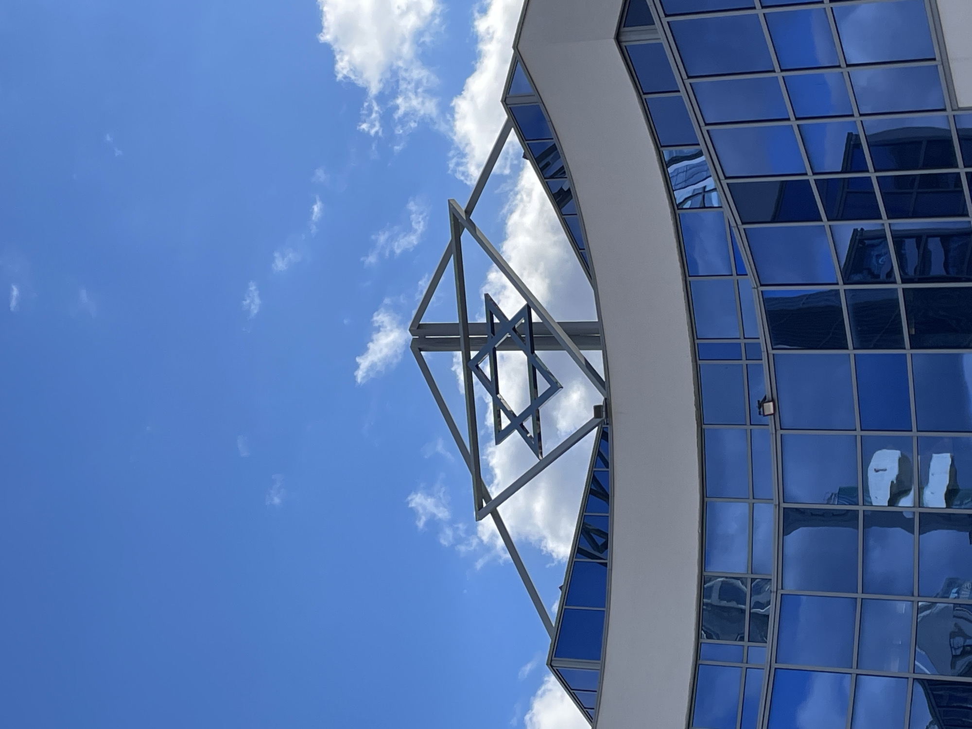 The image shows a portion of a building in Yekaterinburg, Russia. The building has a curved facade with blue-tinted windows. A metal structure, possibly an architectural detail, extends from the building against a blue sky with scattered clouds. The windows reflect the sky and surrounding buildings. A spotlight is affixed to the building's exterior. The image is oriented so that the building is positioned on its side, with the sky occupying the majority of the frame.
