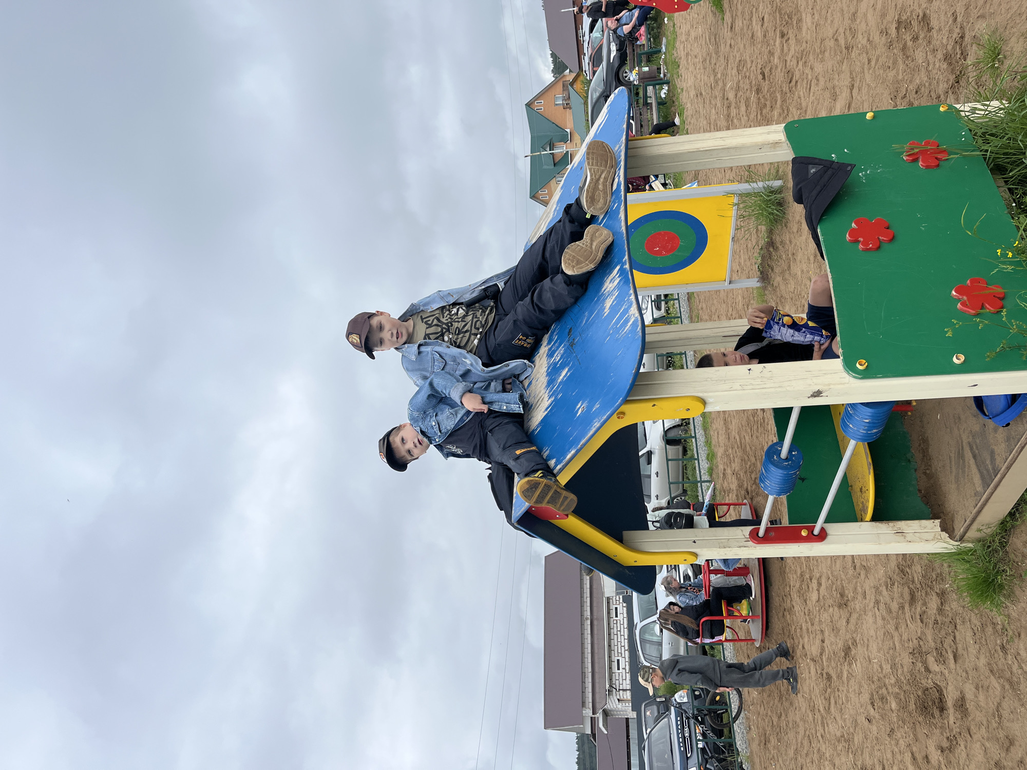 This image captures an outdoor playground scene in an unknown residential area under an overcast sky. The ground consists of sandy dirt with patches of green grass. In the foreground, a multi-colored playhouse features a weathered blue roof, yellow supports, and green side panels adorned with three red flower cutouts and two horizontal bars with blue cylindrical weights.
Two pre-teen boys are sitting on the blue roof of the playhouse. The boy on the left wears a brown baseball cap, a denim jacket over a dark t-shirt, dark pants, and shoes with yellow soles; he looks towards the right. The boy on the right, appearing slightly older, wears a brown baseball cap, a denim jacket over a camouflage-patterned t-shirt, dark pants, and tan athletic shoes; he looks towards the left with his legs extended. Inside the playhouse, a younger boy in a dark t-shirt and shorts sits on the floor, holding a bag of snacks.
To the left of the playhouse, a red merry-go-round is visible, with three pre-teen girls sitting on it. One girl has long dark hair, another has lighter hair, and the third is partially obscured. Near the merry-go-round, a pre-teen boy in a grey tracksuit and a camouflage baseball cap stands next to a bicycle. In the mid-ground, behind the playhouse, a yellow rectangular board with a red, green, and blue target symbol stands upright.
The background reveals several parked cars, including a dark Kia Sportage, and multiple houses with varying architectural styles, some featuring brown roofs and light-colored walls, and another with a green roof. Several adults are present in the background, some sitting on a green bench, others standing near the vehicles. Overhead, electrical wires traverse the cloudy, grey sky.