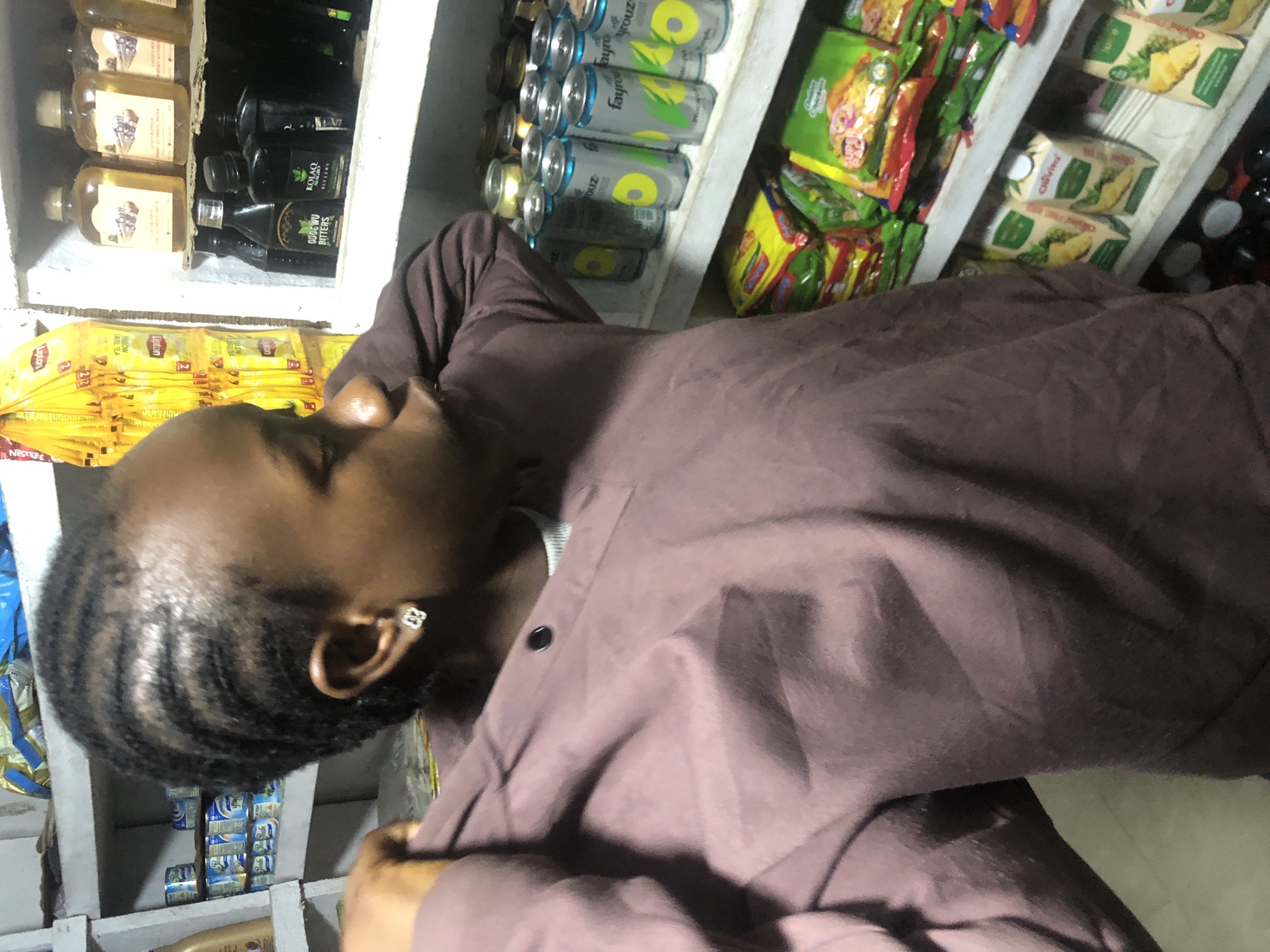 The image shows a young Black woman in a dimly lit shop, possibly a small convenience store or local market in Ado, Nigeria. She is wearing a muted purple sweatshirt and a white tank top underneath. Her hair is styled in braids, and she has a small, square earring in her left ear. She is looking to her right and her left hand is pulling at the sleeve of her sweatshirt. The background is filled with shelves stocked with various products. On the top shelf, there are bottles of what appear to be alcoholic beverages and some packaged goods. The middle shelf holds several cans of a beverage, some of which are labeled "Fayrouz". The bottom shelf displays packets of snacks and juice cartons. The overall impression is a candid shot of a person in a commercial environment.