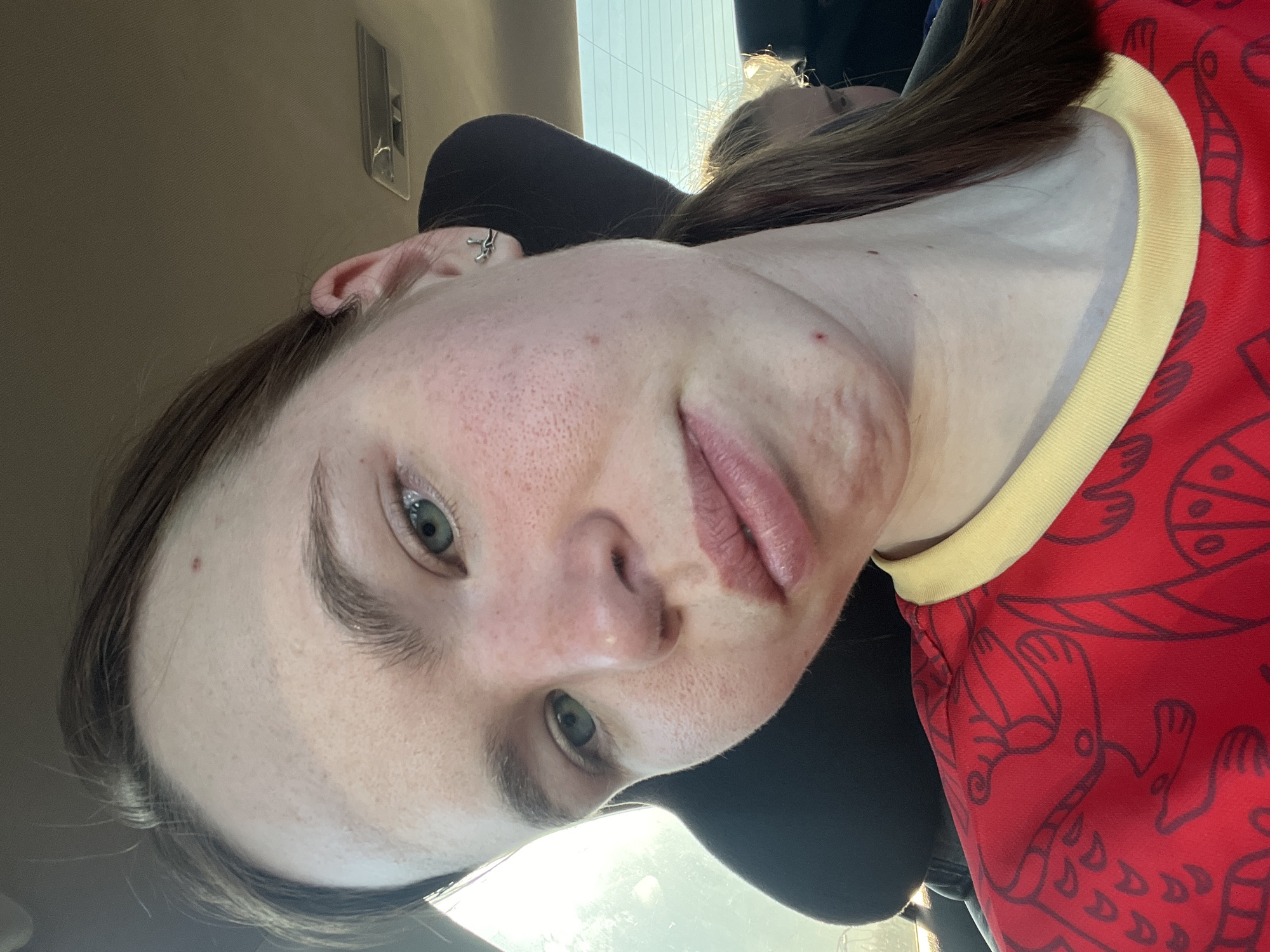 A close-up selfie depicts a young woman with fair skin and light brown hair. She is seated in what appears to be a car, based on the interior elements visible in the background. The woman is wearing a red shirt with a yellow collar and a pattern of dark lines, possibly depicting stylized animal figures. The location is uncertain.