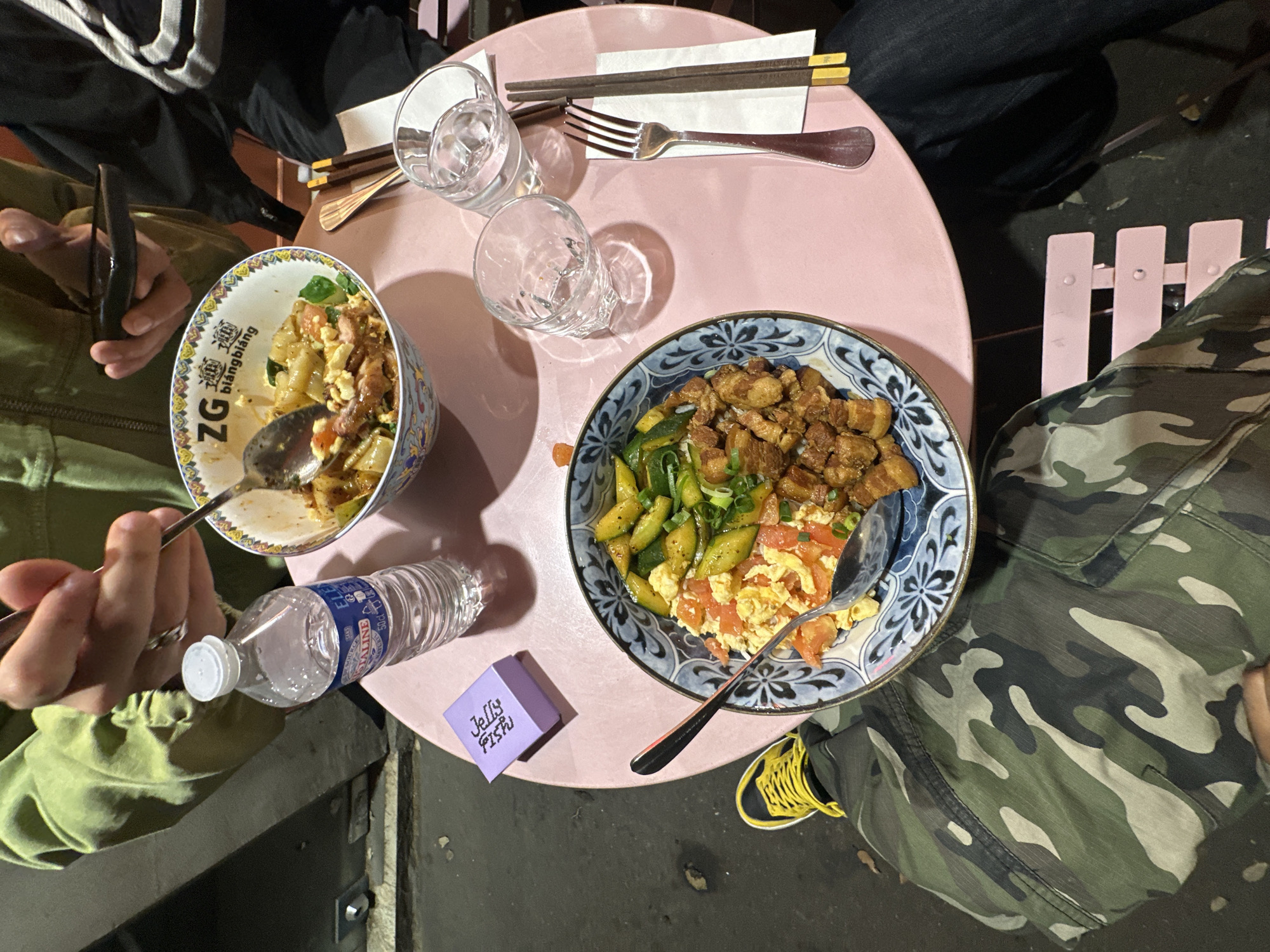 This overhead shot captures a casual evening meal around a round, pink table, likely in an outdoor or semi-outdoor setting. At least three individuals are present, indicated by their visible clothing and hands: one wears a green jacket, another dark clothing, and a third, camouflage pants with yellow-laced shoes.
Two distinct dishes are the central focus. The bowl on the left, adorned with "ZG" and the Chinese characters for "biángbiáng" (biang biang noodles), contains wide noodles, scrambled egg, and other components, being eaten with a spoon. The bowl on the right features diced meat, resembling pork belly, alongside sliced cucumbers, and scrambled egg mixed with tomatoes, also served with a spoon.
The table is set with two clear glasses, one holding water, accompanied by a pair of chopsticks on a paper wrapper ("ZG BIANG BIANG") and a fork. A plastic bottle of "CRISTALINE" water lies on the table, near a small, purple box labeled "Jelly Fish". A person on the left holds a dark-colored phone or device. The ground beneath the table appears to be dark concrete or asphalt, and parts of pink slatted chairs are visible.
The artificial lighting strongly suggests the meal is taking place during the evening or night. The presence of Cristaline water, a common French brand, supports the provided location context of Paris, France, pointing to a relaxed dining experience in the city.
