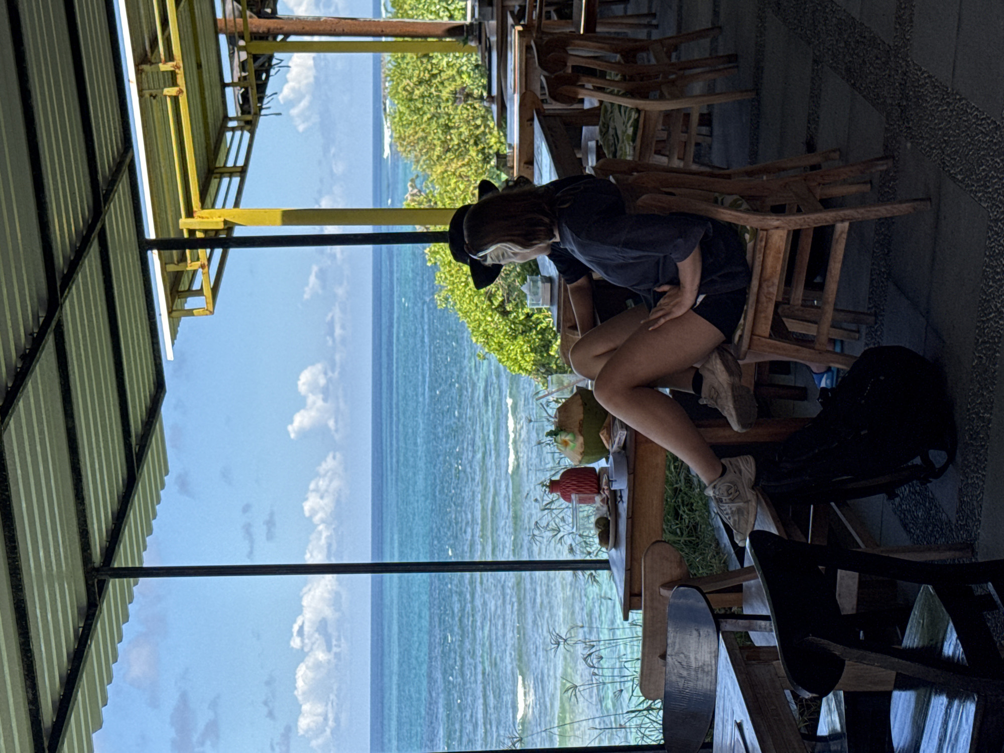 This image captures a bright, daylight scene at an outdoor or semi-outdoor cafe or restaurant overlooking the ocean in Kuta Selatan, Indonesia.
The primary subject is a young person, likely female, with short dark hair, wearing a dark top, dark shorts, a dark hat, and light-colored sneakers. She is seated in a wooden chair at a rustic wooden table, with her hands resting casually on her lap/thigh. On the table in front of her, a green coconut drink with a straw and a red bottle or container are visible.
The establishment features a simple, corrugated metal roof supported by black and yellow metal beams, providing shade for the diners. Beyond the seating area, a band of vibrant green foliage separates the cafe from the vast, clear turquoise ocean, which stretches out under a bright blue sky adorned with scattered white clouds. Gentle waves are visible on the water's surface. Other wooden chairs are seen in the background, some stacked.
The scene evokes a sense of relaxation and leisure, typical of a tropical coastal setting. The weather appears sunny and pleasant. No visible text is present in the image.