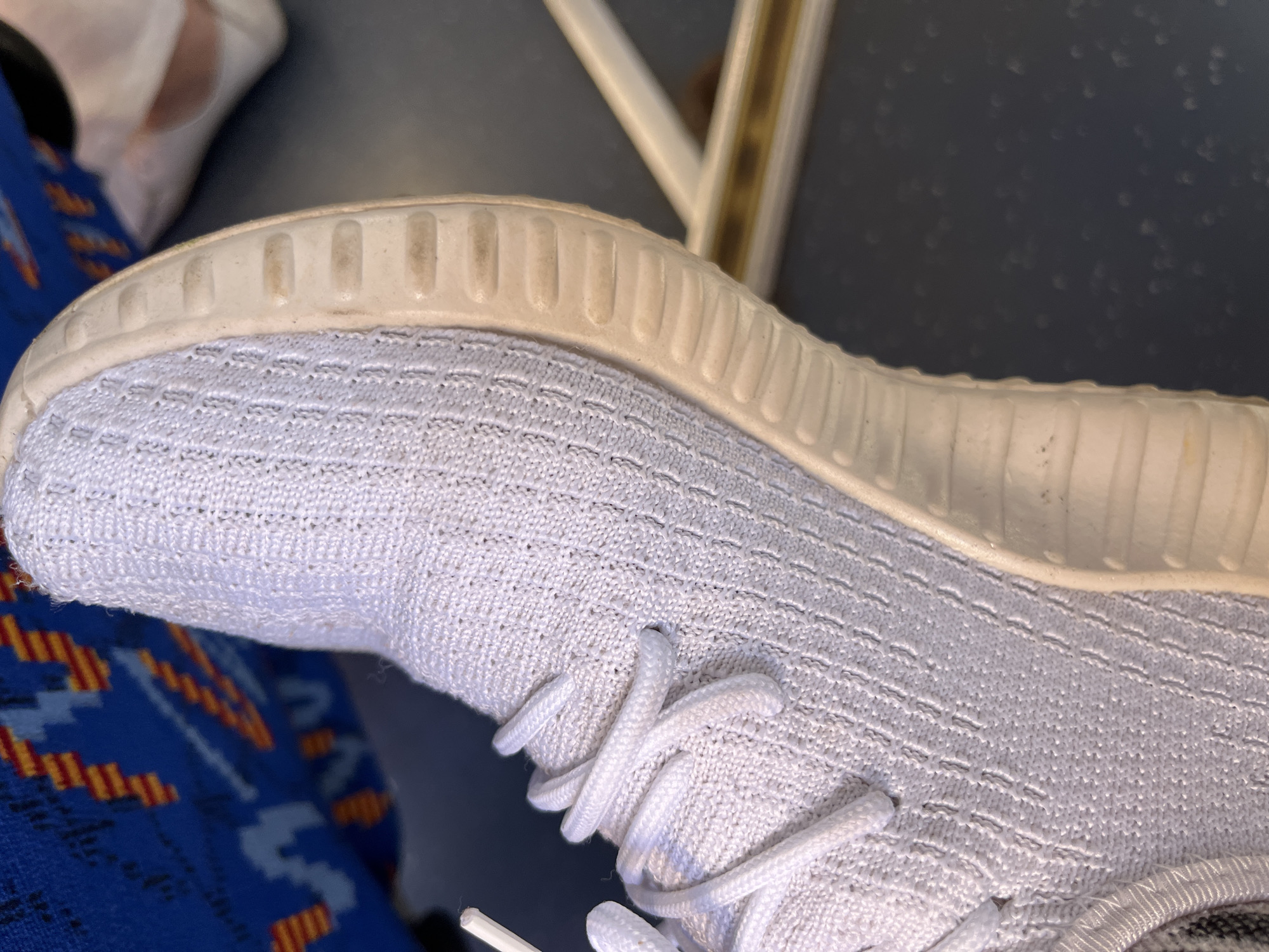 The image shows a close-up of a white knit sneaker with white laces. The sole of the shoe is light beige and has a textured pattern. The shoe is resting on a dark surface. In the background, out of focus, a blue and orange patterned fabric is visible, possibly clothing. Another shoe, partially visible and lighter in color, is also in the background. The environment appears to be indoors, possibly on a public transport. The scene is devoid of any specific Myadzyel landmarks.