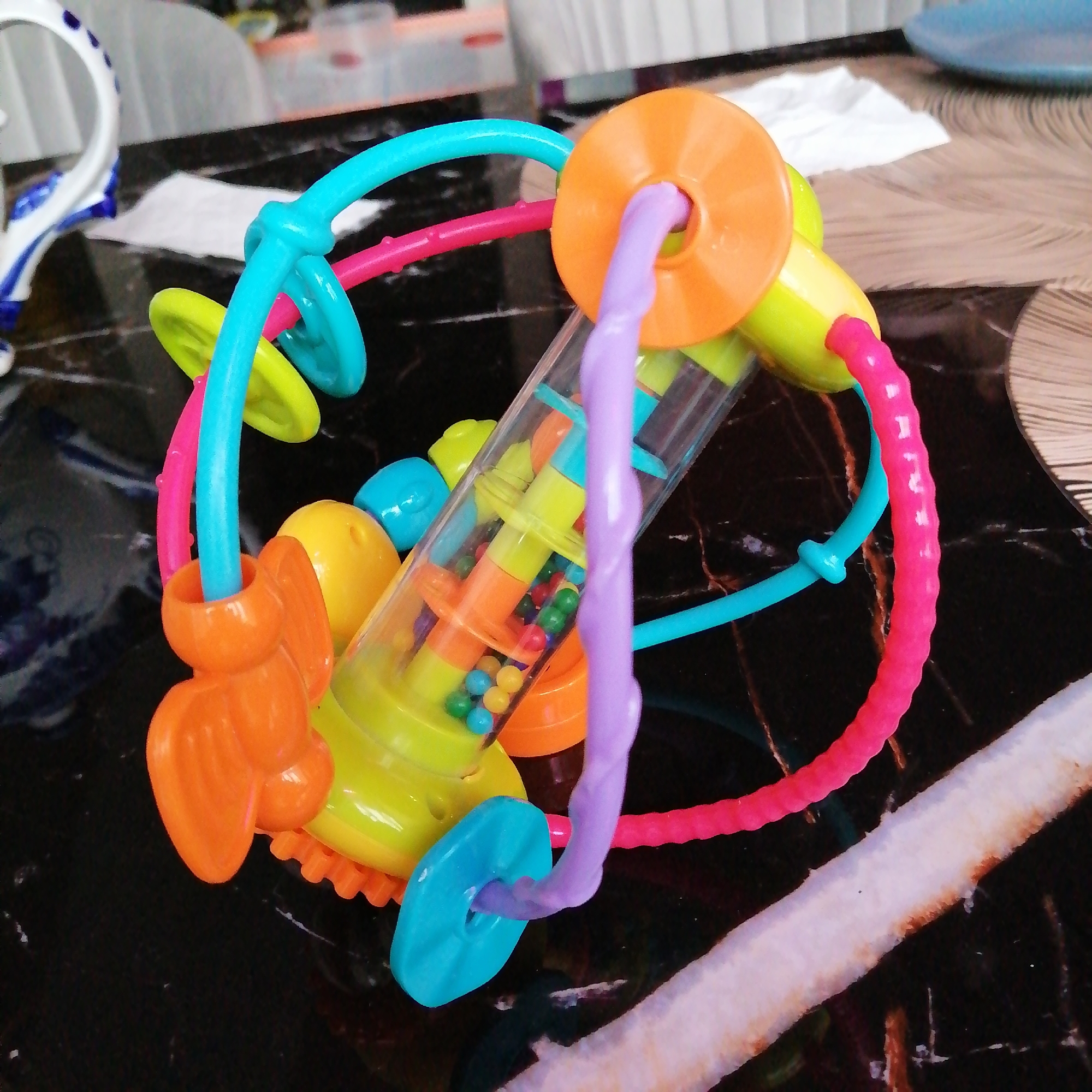 Here's a detailed description of the image:
**Scene:** An indoor setting, likely a home, on a surface with a black and gold speckled pattern.
**Object:** A brightly colored baby toy dominates the scene. The toy is spherical in shape with various colorful, rounded and angular elements. There are loops and beads incorporated into the design. A clear plastic tube containing small, colorful balls runs through the center of the toy. The colors are vibrant and include turquoise, pink, orange, yellow, and purple.
**Other Details:**
* A white cloth napkin is visible in the background.
* A partially visible white object with blue trim is to the left.
* A brown/tan object is on the right side of the toy.
**Location:** The precise location is unknown, but given the objects present, it is likely a domestic environment, such as a kitchen or dining area.