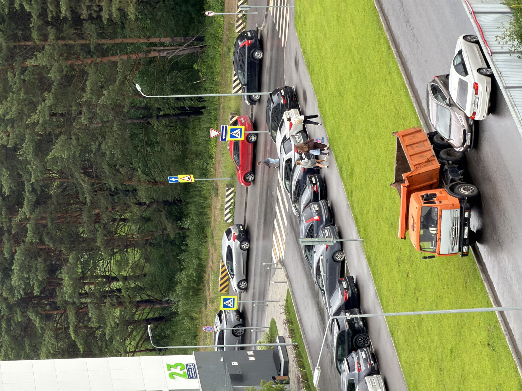This is an aerial photograph of a street with several cars parked along the side. The scene appears to be a driving school or a similar training facility, as indicated by the presence of traffic cones, road signs, and a marked driving course.
In the foreground, an orange dump truck is parked, its bed raised, suggesting it might be involved in construction or landscaping. Several cars are parked in a line along a wet asphalt road, indicating it may have recently rained. A group of people are standing near some of the parked cars, some appearing to be students or instructors.
There are several road signs visible, including a pedestrian crossing sign and a directional sign. The trees lining the road are lush and green, suggesting a temperate climate. The sky is overcast, indicating a cloudy day. The overall atmosphere appears to be one of activity and learning, with the presence of both vehicles and people engaged in various tasks.
On the bottom left of the image, there is a white building with the number "23" in green text and some other text which is difficult to read. Some license plates are visible on the cars, but the text is not legible. The time of day is difficult to determine precisely, but the lighting suggests it is daytime.