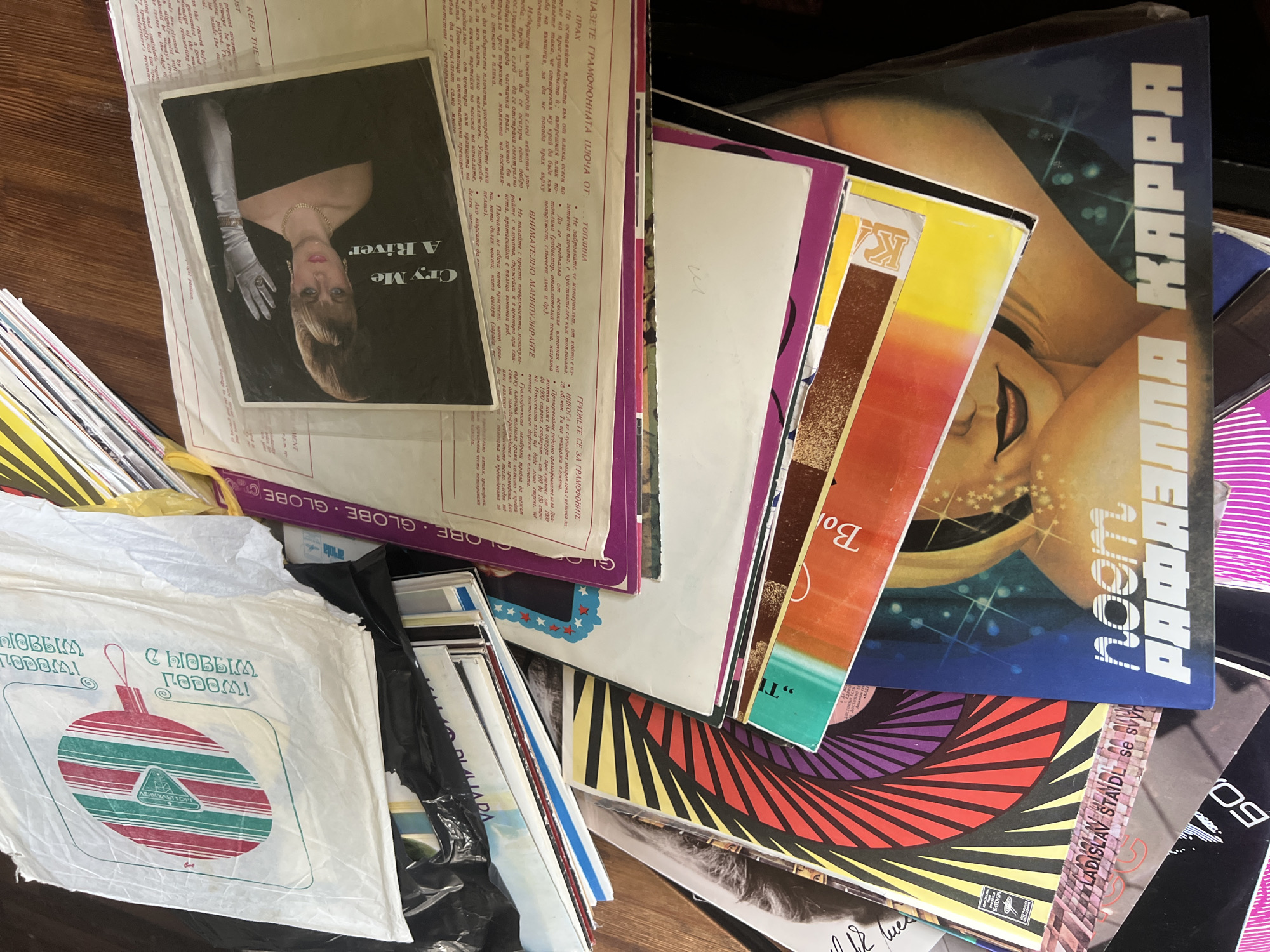 The image shows a pile of vinyl records and their sleeves on a wooden surface. The records are of various sizes and colors, with some having prominent graphic designs. One record sleeve features a close-up of a woman wearing white gloves and jewelry, with the text "Cry Me A River" visible. Another record sleeve displays a colorful abstract design with geometric shapes in red, yellow, black, and purple. Further down, a record cover features a portrait of a woman with the text "noem РАФАЭЛЛА КАРРА". There are also several white plastic bags, one of which has a printed design of a red and green striped ornament and Cyrillic text. The overall scene suggests a collection of music records, possibly in a storage or display setting. The provided context does not specify a location in Ленинградская, Russia.