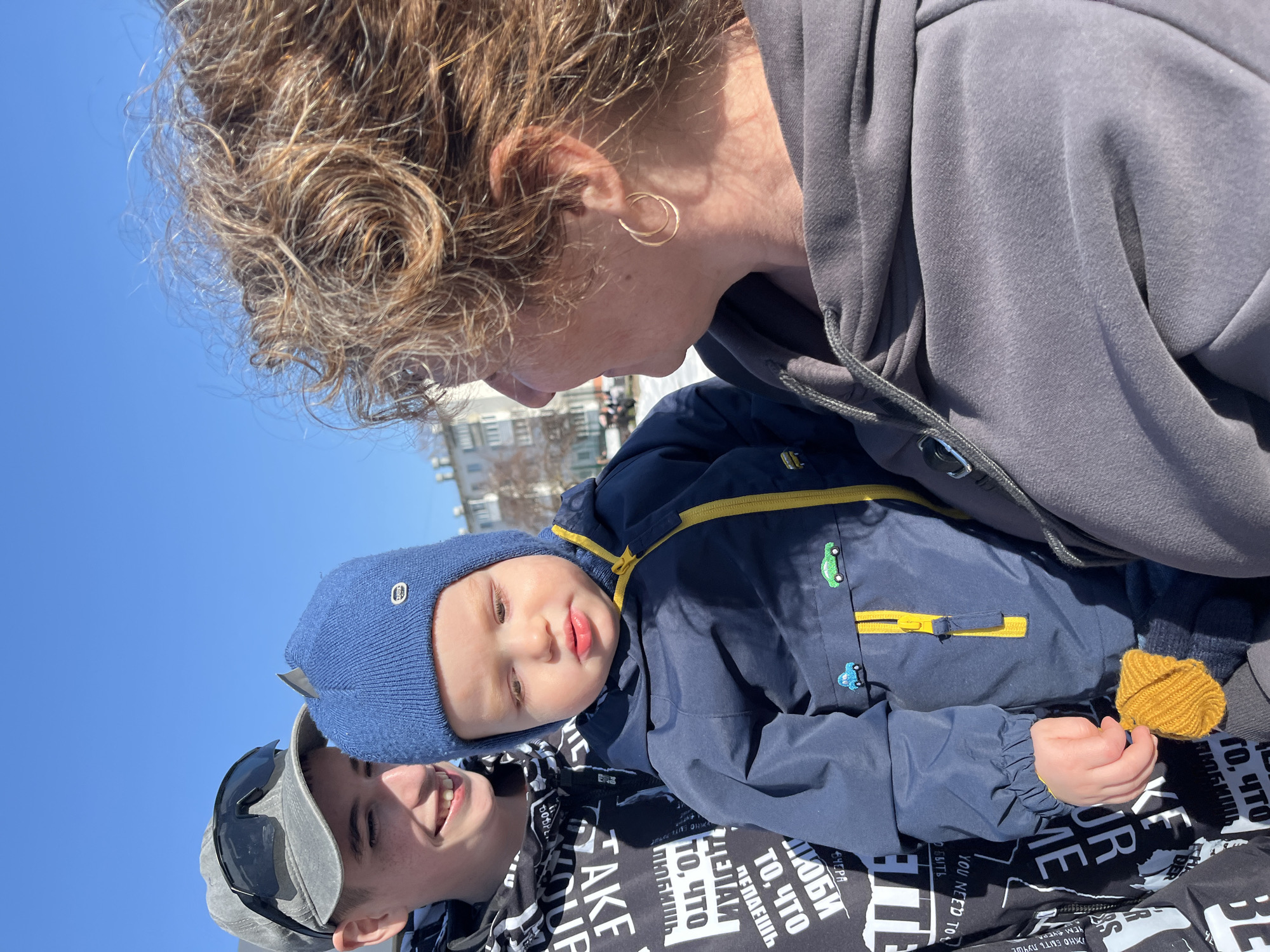 The image shows three people in an outdoor setting. A woman with curly hair, wearing a gray hooded sweatshirt and gold hoop earrings, is looking down at a young child in a blue hat and jacket with yellow details. The child is holding a yellow knitted object. The third person, a young man wearing a gray cap and sunglasses, is partially visible at the bottom of the frame.
The background includes a clear blue sky and a building in the distance. The clothing of the young man underneath the child is black and white with Russian lettering. Based on the presence of Cyrillic text, and the request for the location, it is reasonable to identify the location as the city of Chaikovsky, Russia.