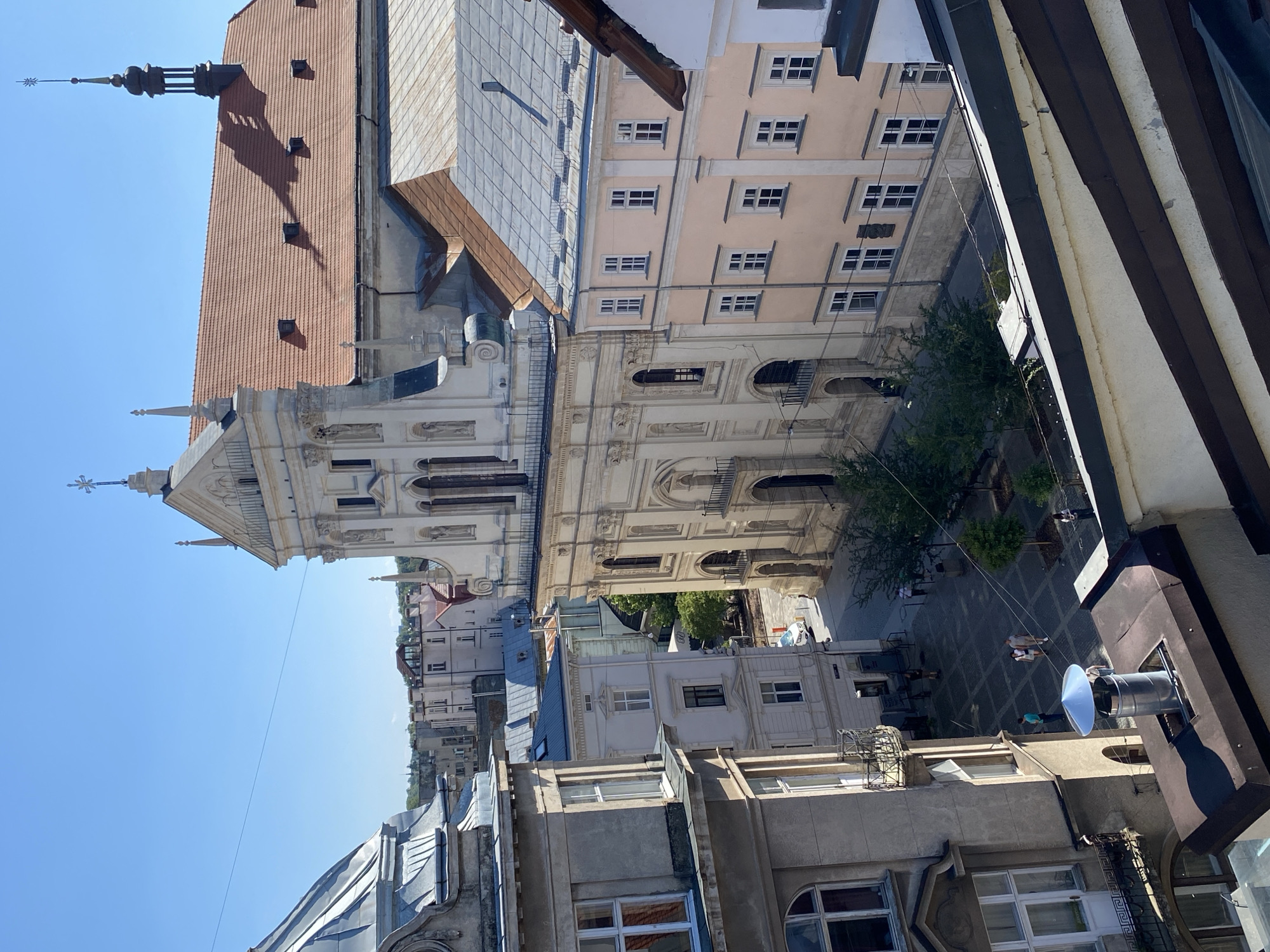 The image shows a view of a street in Lviv, Ukraine, with several historic buildings. In the foreground, a metallic chimney vent protrudes from a dark, angled roof. To the left, a light-colored building with multiple windows, balconies, and architectural detailing is visible.
Dominating the center and right of the image is a large, ornate white building with a classic facade, featuring columns, sculptures, and arched windows. It has a terracotta-tiled roof and a tall, dark spire with a cross on top. Adjacent to this building, to its right, is a pale pink building with rows of white-framed windows.
Below the buildings, a cobblestone square is visible with scattered trees and a few people walking. A white car is parked on the street. The sky is clear blue. The overall impression is of an urban historical district bathed in sunlight.