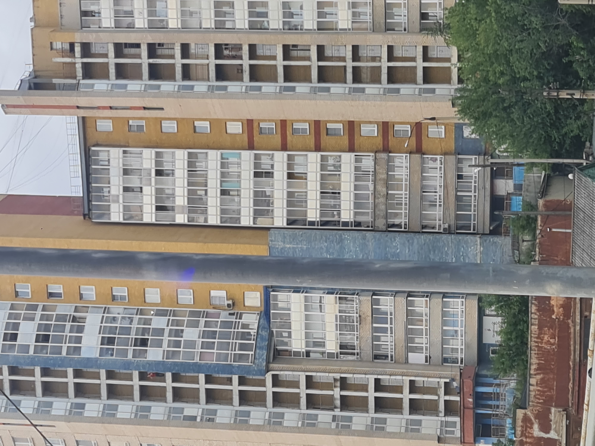 The image captures a segment of a tall, multi-story residential building in Irkutsk, Russia. The building features a mix of architectural styles and colors, with sections clad in beige, yellow, and blue materials. The facade is characterized by numerous windows and balconies, some enclosed and others open. Several levels of balconies are visible, many with white framing and clear glass. The building appears to be an apartment complex with multiple units.
In the foreground, a thick, grey metal pole, possibly a lamppost or part of a utility structure, partially obscures the building. The lower portion of the image reveals a rusty metal barrier or wall, suggesting a construction site or an industrial area adjacent to the residential building. Behind this barrier, dense green foliage, likely trees and bushes, forms a natural screen, partially concealing lower structures and possibly a road or path.
The overall environment suggests an urban setting in Irkutsk, Russia, with the residential building being a prominent feature. The presence of trees and the rusty barrier indicates a contrast between modern urban development and remnants of older infrastructure or industrial activity. The sky is overcast, contributing to a subdued lighting condition. There are no discernible individuals engaged in specific actions or interactions, but a person in a red shirt can be seen on a balcony in the upper left section of the building. The scene is static and observational, presenting a snapshot of the urban landscape.